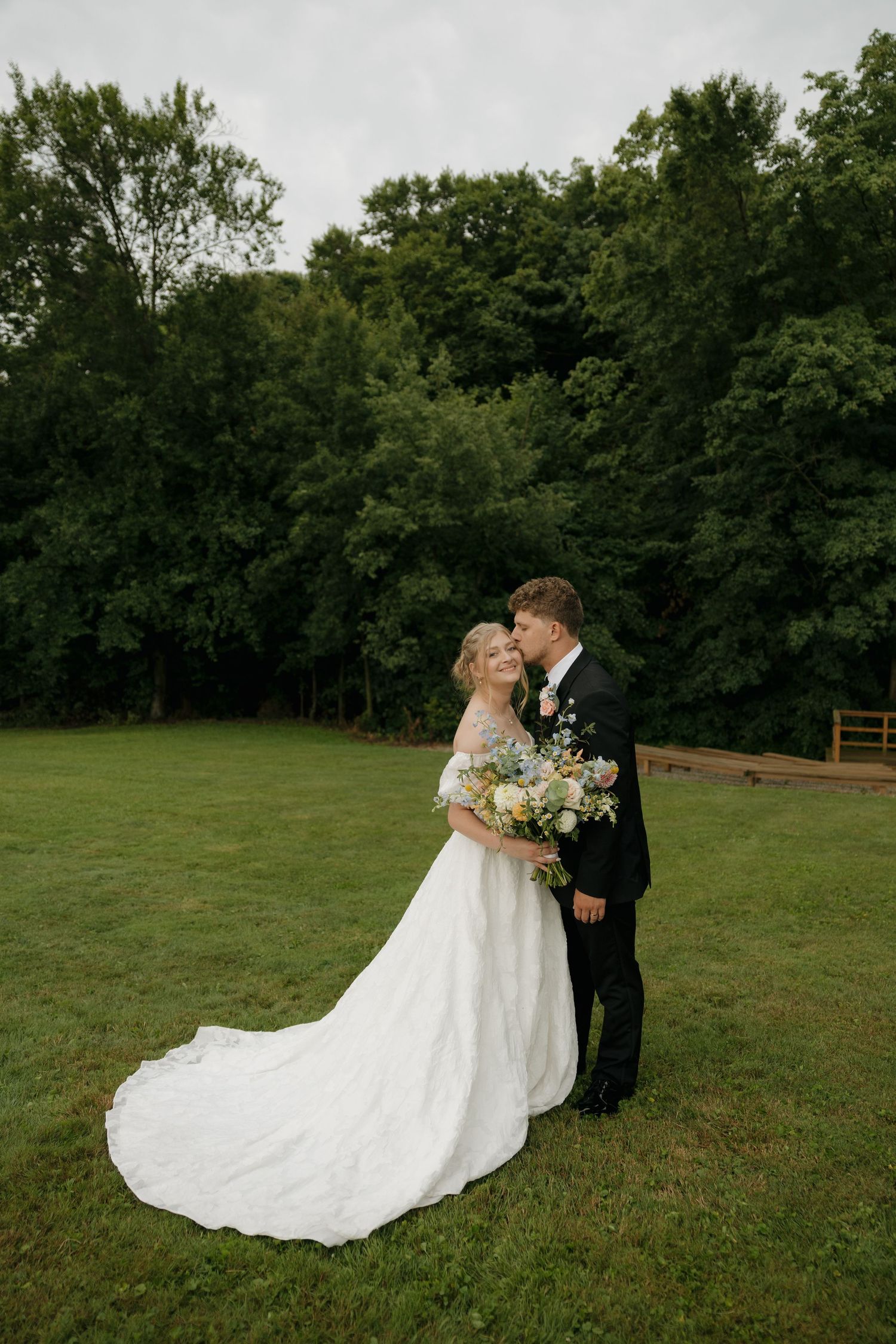 Bride and groom share a romantic moment on a lush green lawn surrounded by trees in an outdoor wedding portrait.