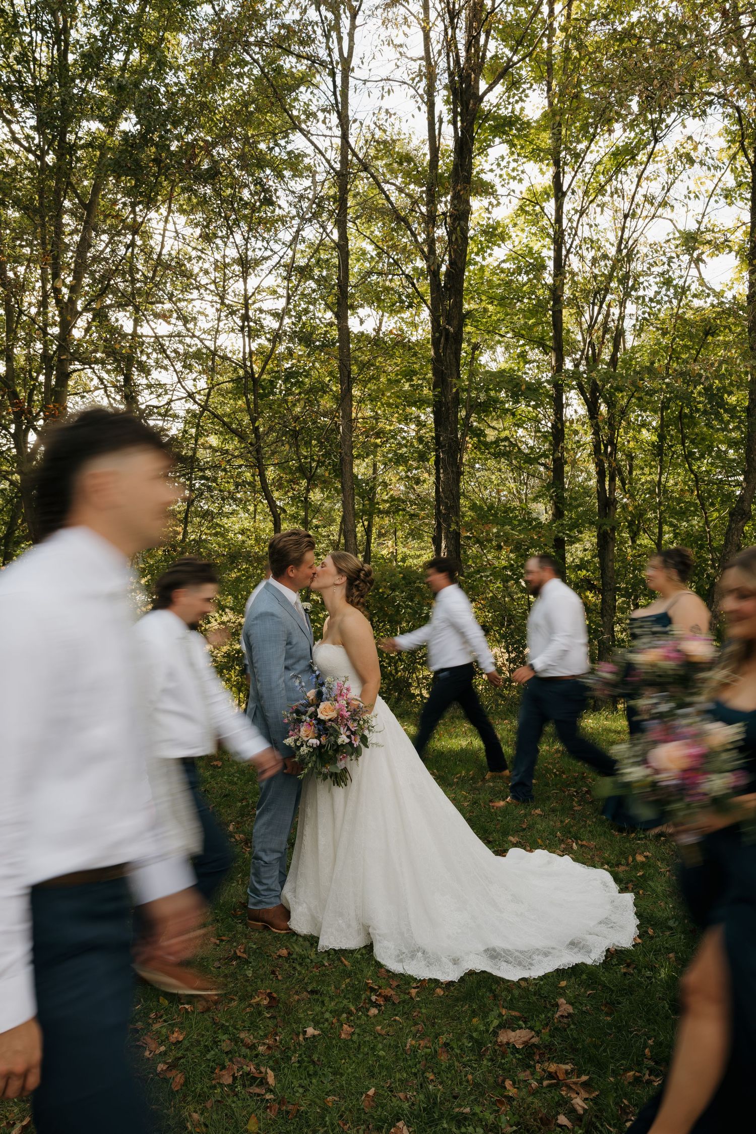 Bride and groom share a kiss in a sunlit forest while blurred wedding guests walk past carrying bouquets.