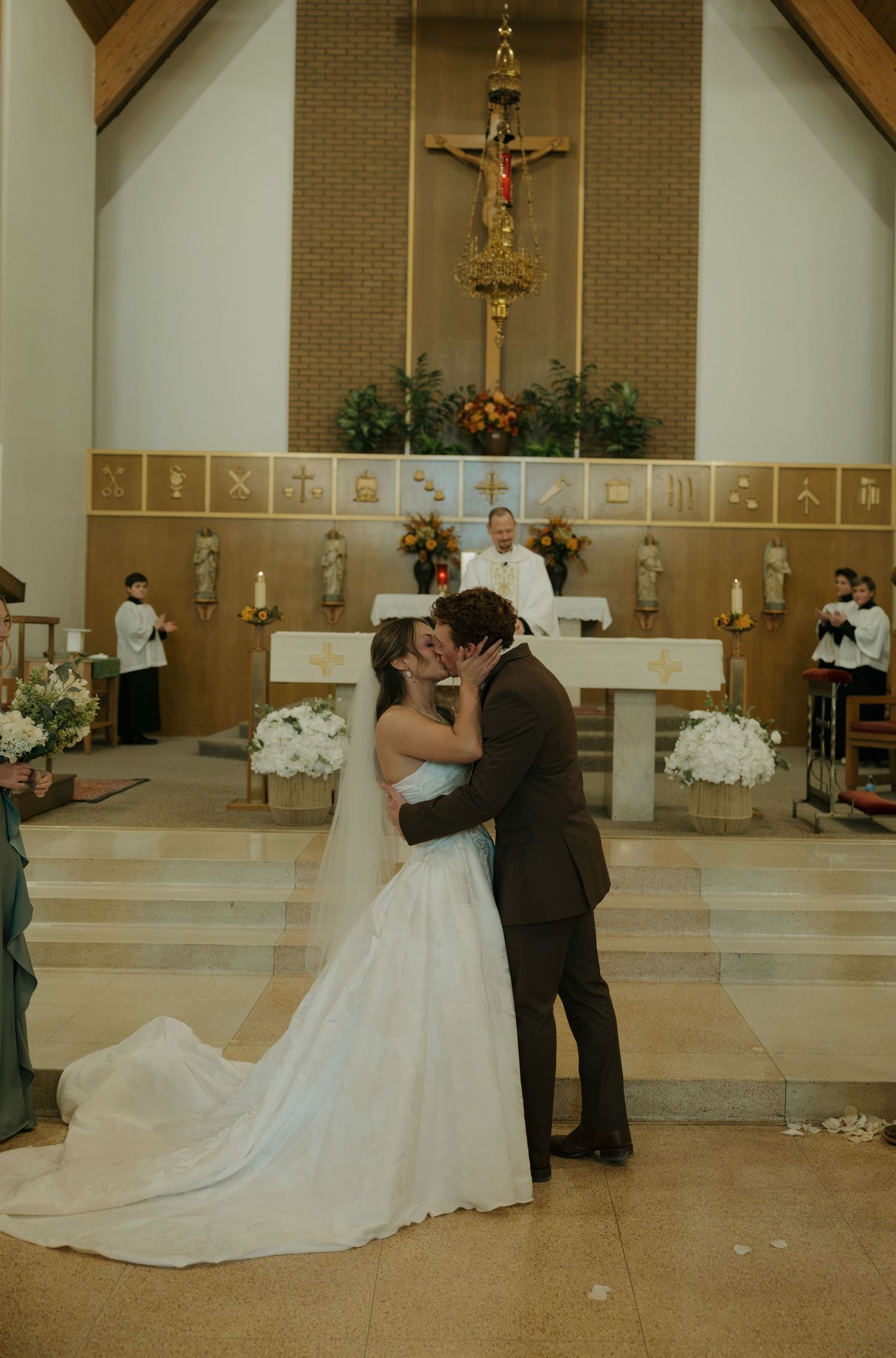Bride in a white gown and groom in a brown suit share a wedding kiss before a church altar with a crucifix, flowers, and priest.