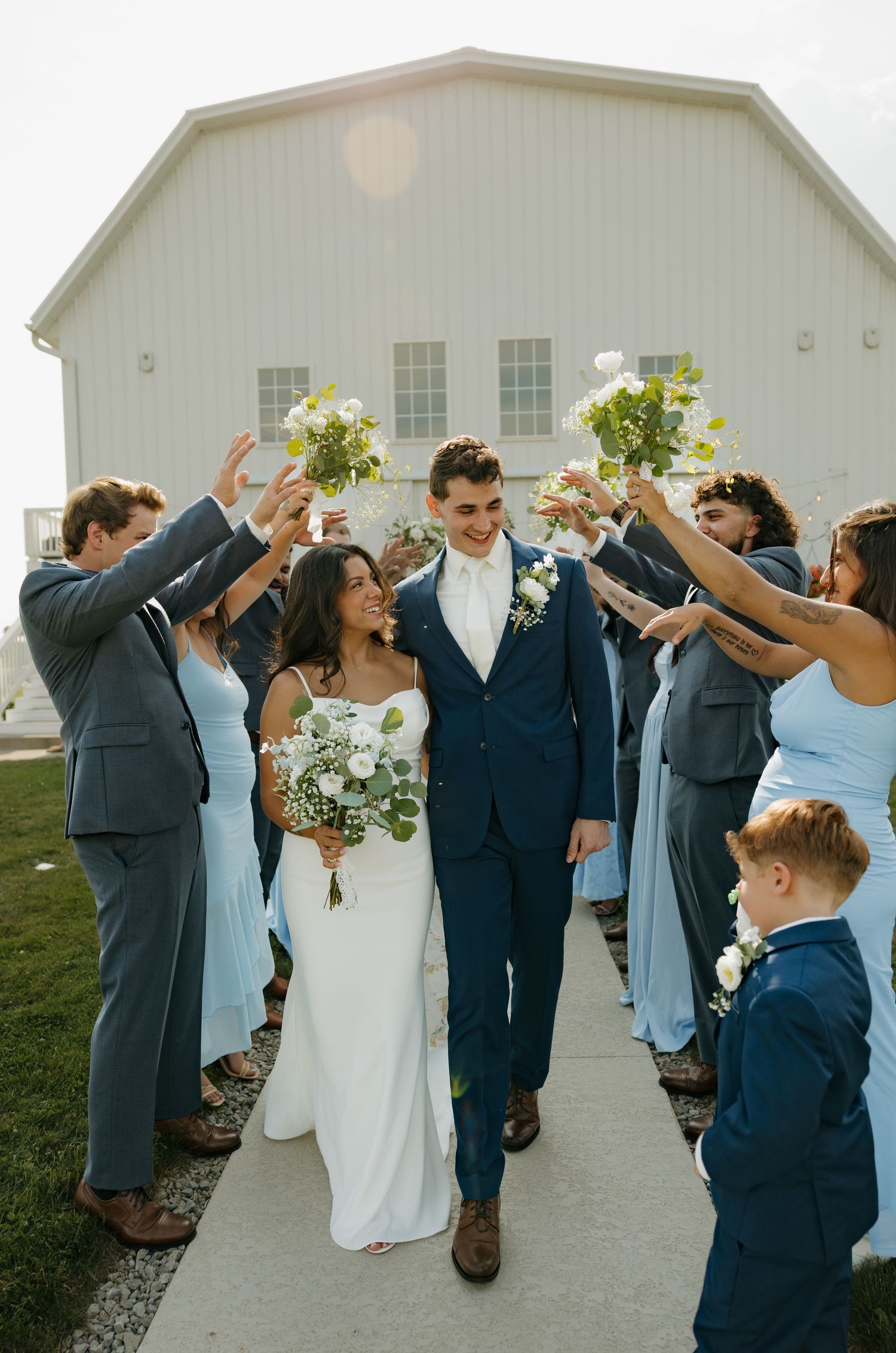 Newlywed couple walks through cheering wedding party outside white barn venue, bride in white gown, groom in navy suit.