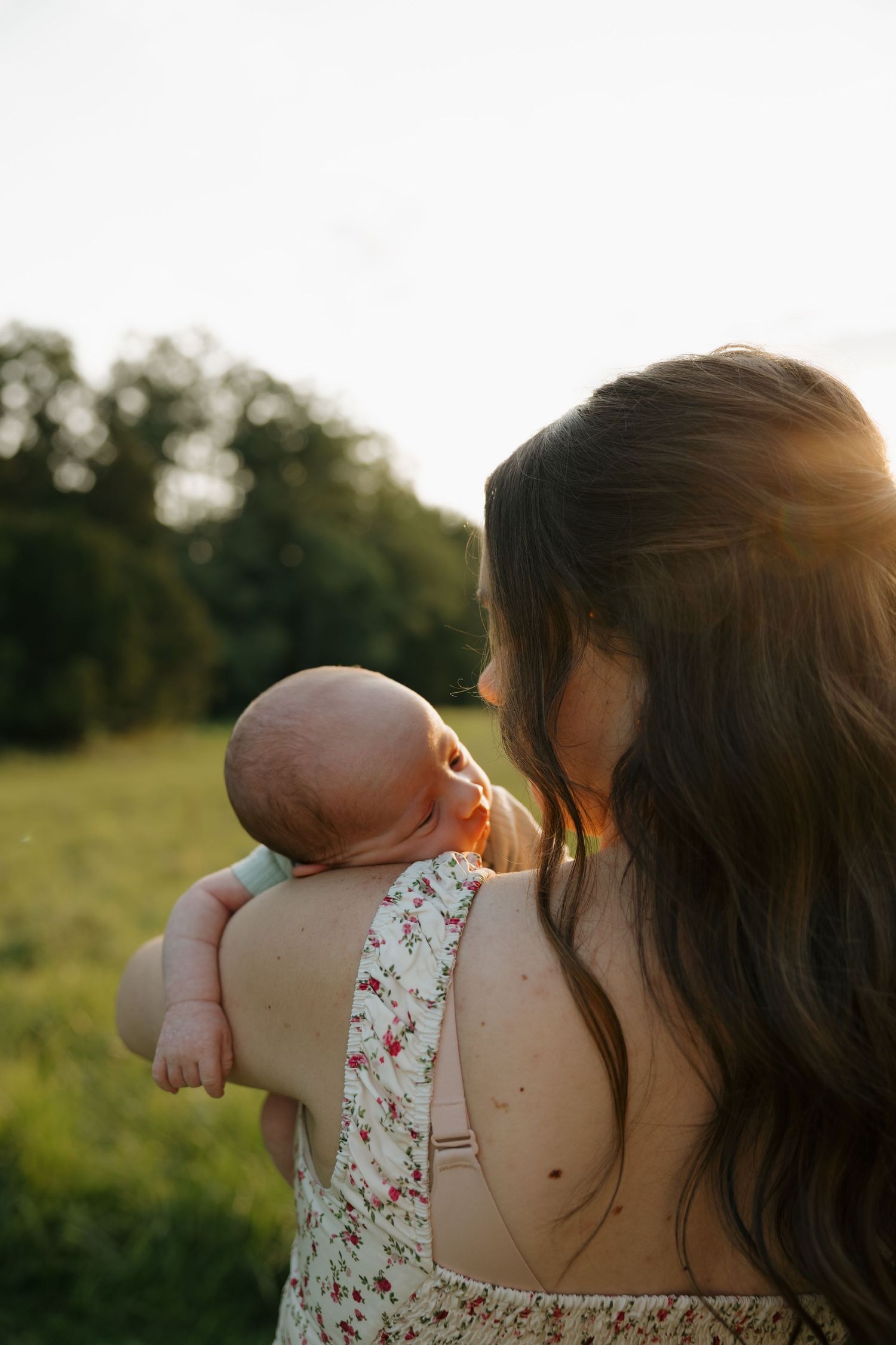 Mother holding newborn baby on her shoulder in a sunlit green field during golden hour, viewed from behind.