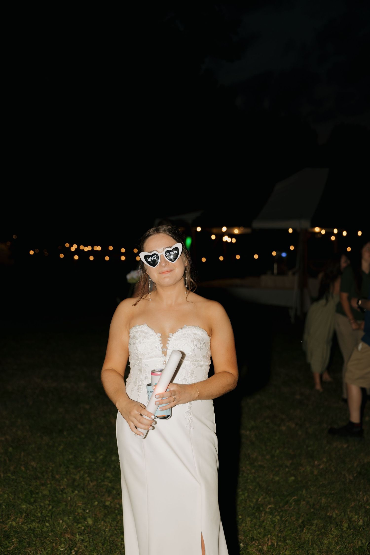 Bride in white strapless gown wearing heart-shaped sunglasses holding champagne at outdoor evening wedding reception.
