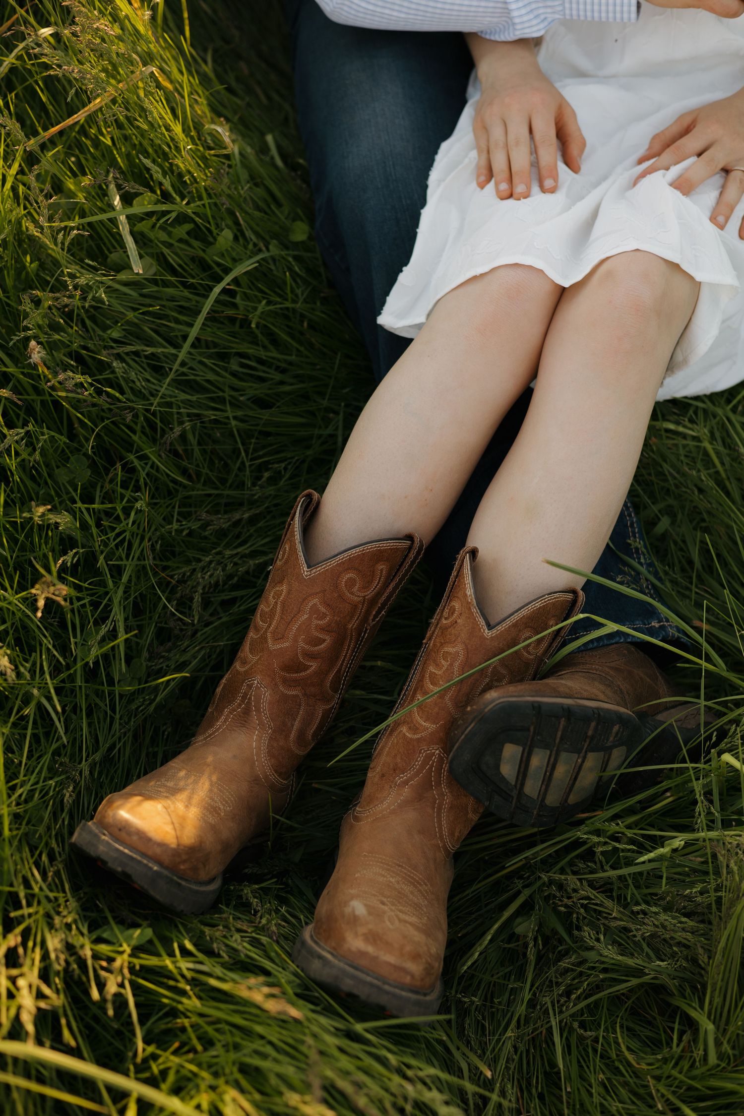 Woman in white dress and brown cowboy boots sitting in lush green grass with a partner beside her.