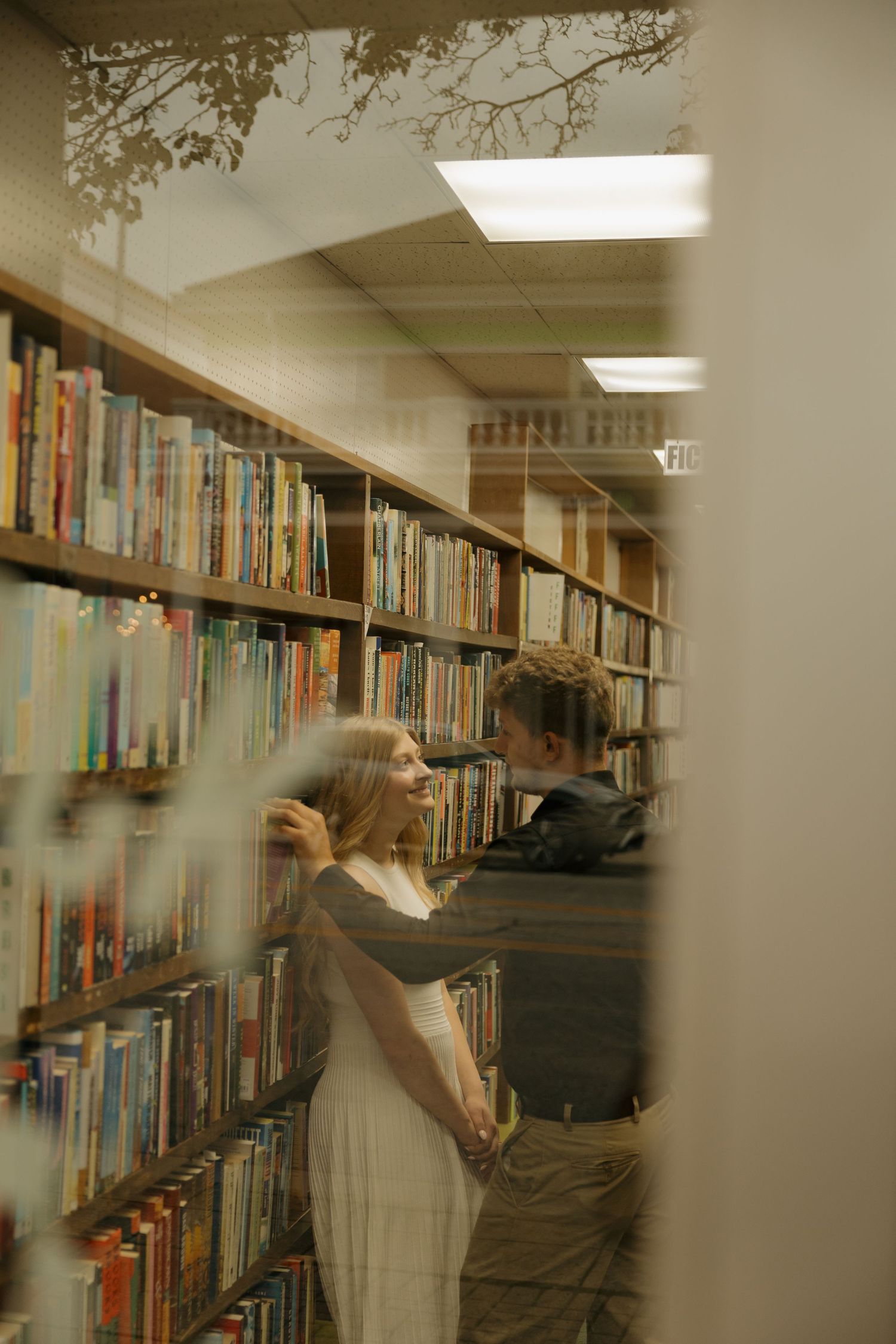 Couple sharing a romantic moment between library bookshelves, photographed through a window reflection.