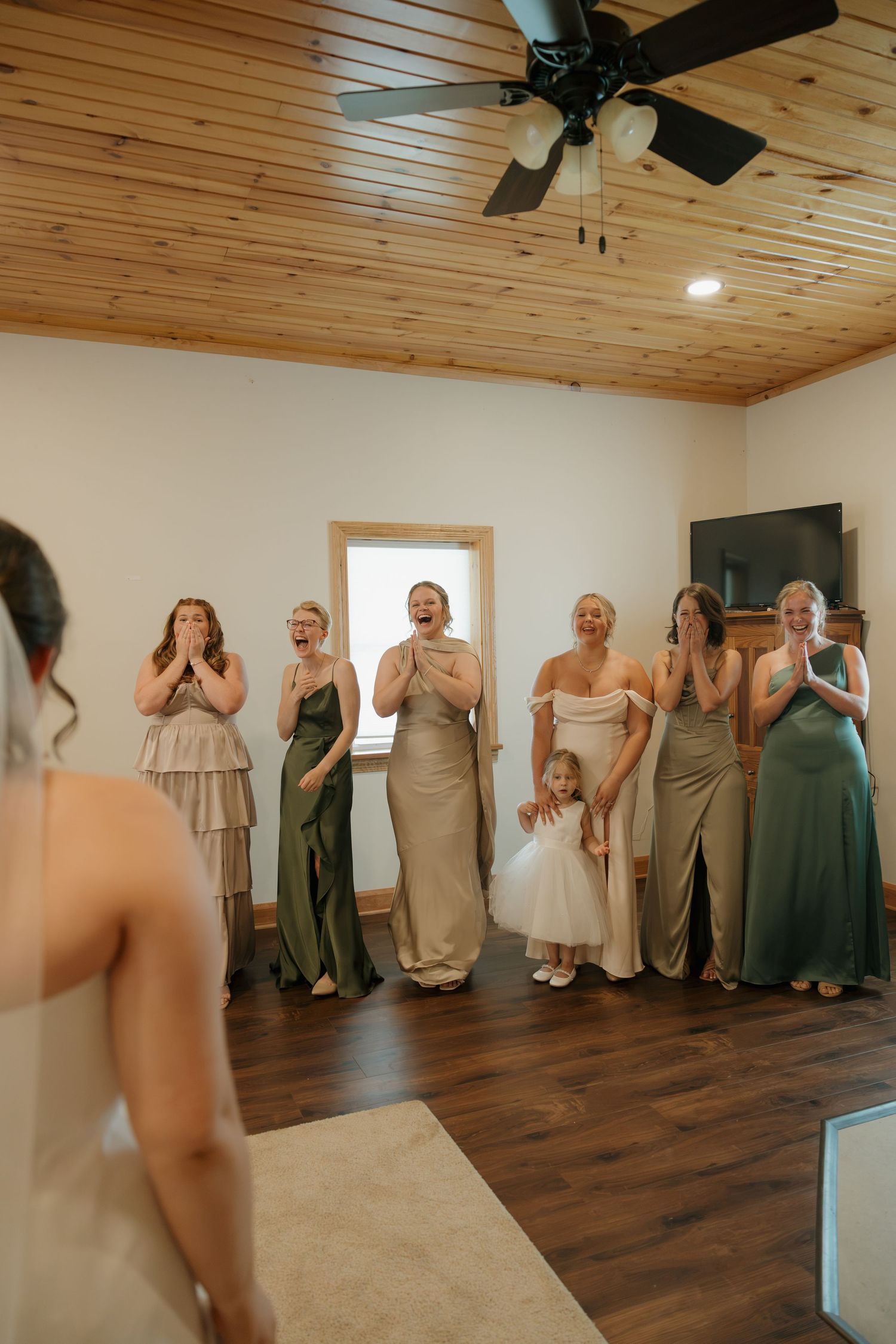 Bridesmaids in sage green dresses react with joy and tears during a wedding dress reveal in a rustic venue.