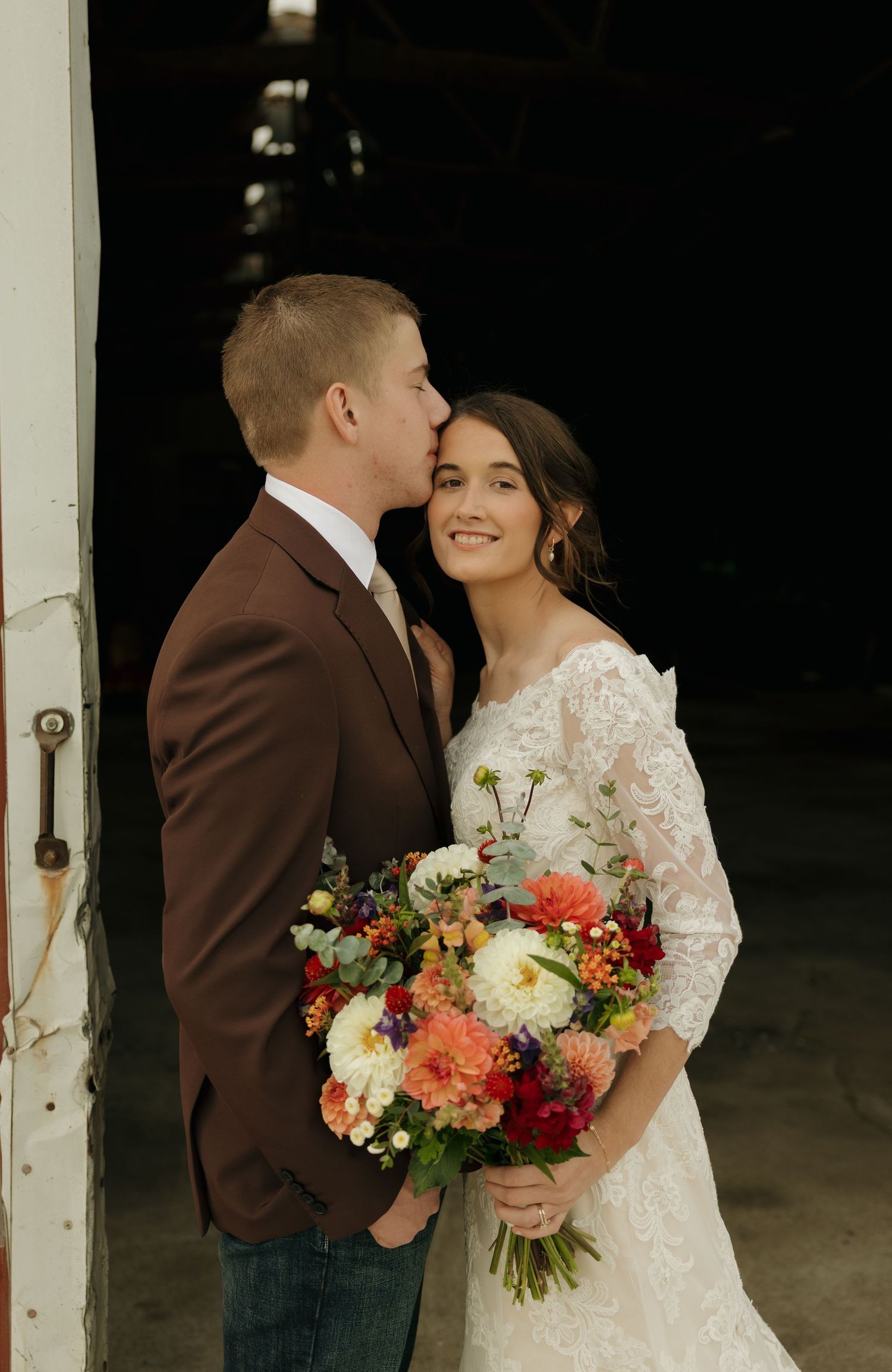 Groom in a brown blazer kisses smiling bride in a lace gown on the forehead as she holds a colorful fall wedding bouquet.