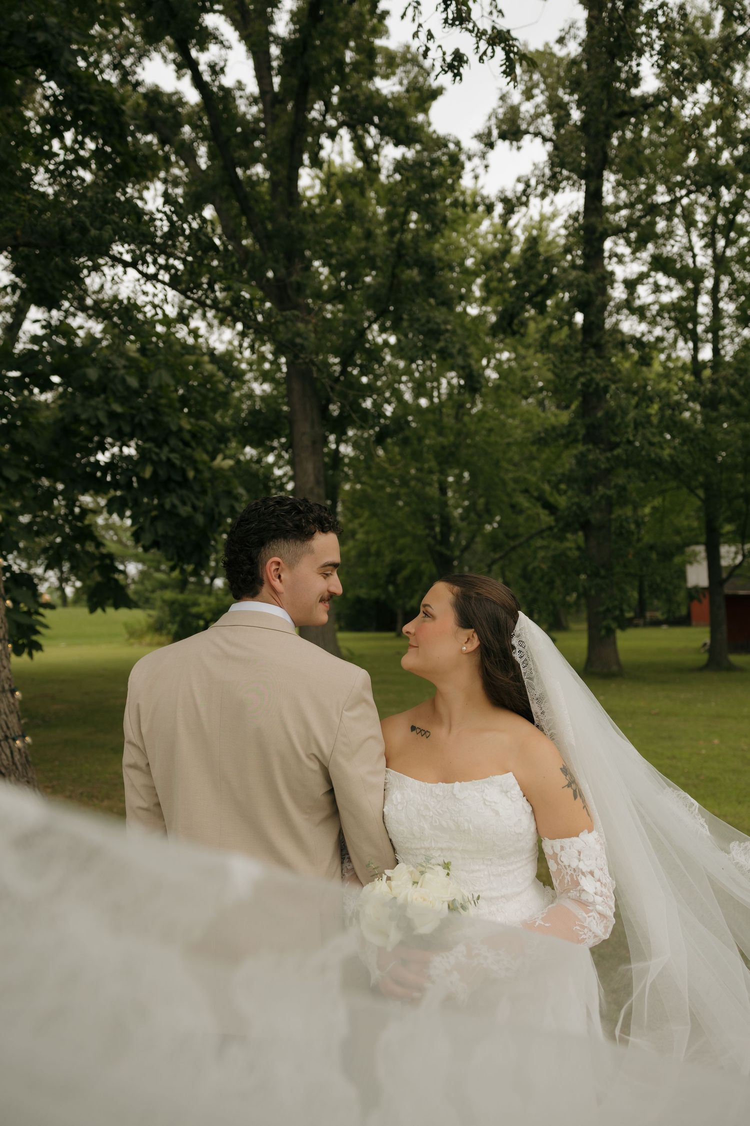 Bride in white off-shoulder gown with veil and groom in tan suit share a romantic glance outdoors.