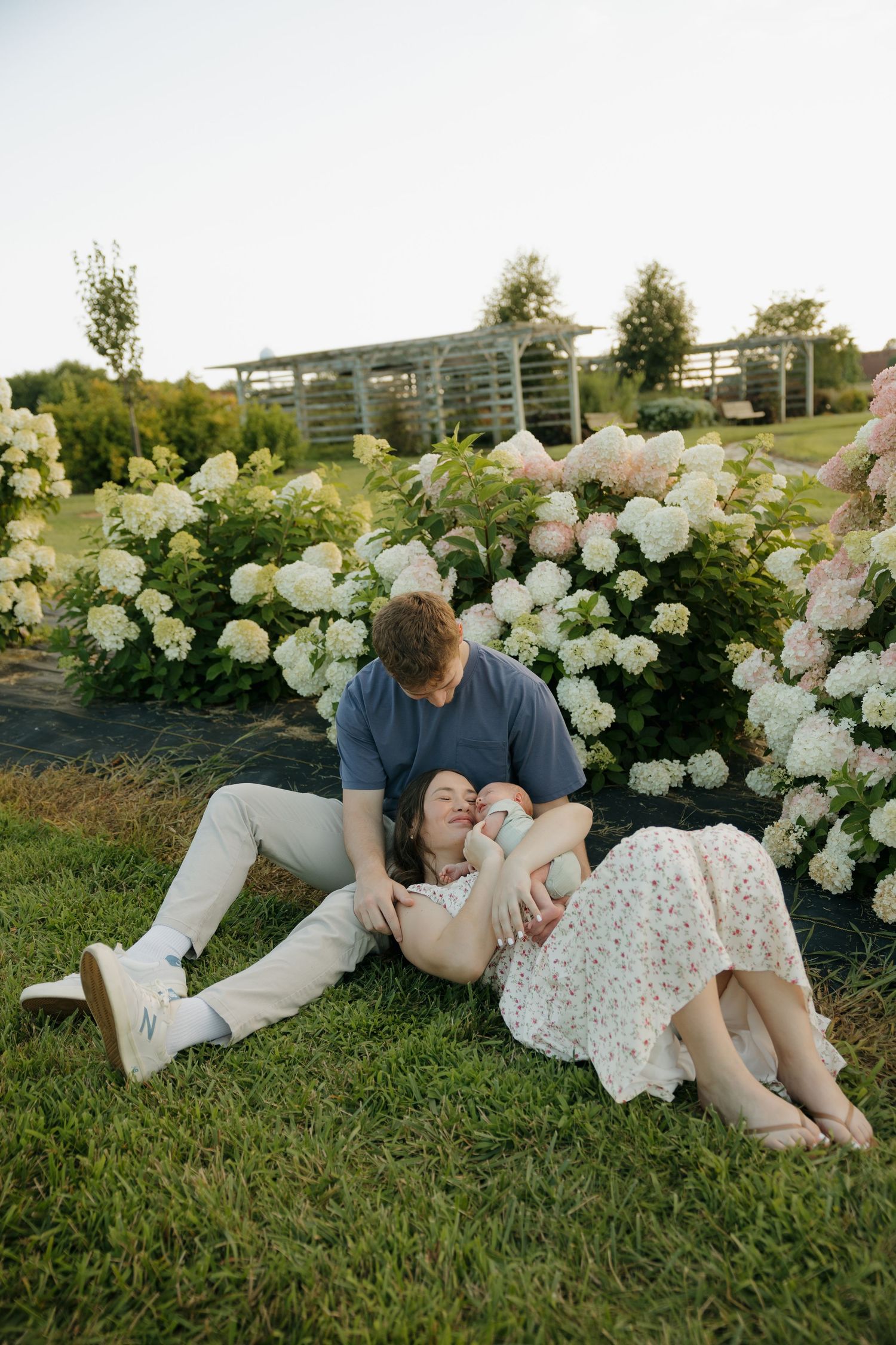 Couple relaxing on grass with newborn baby surrounded by white hydrangea bushes in a garden setting.