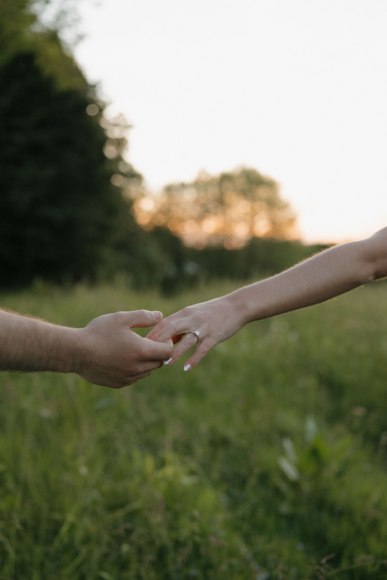Two hands gently touching fingertips in a green field at sunset, with an engagement ring visible on the woman's hand.