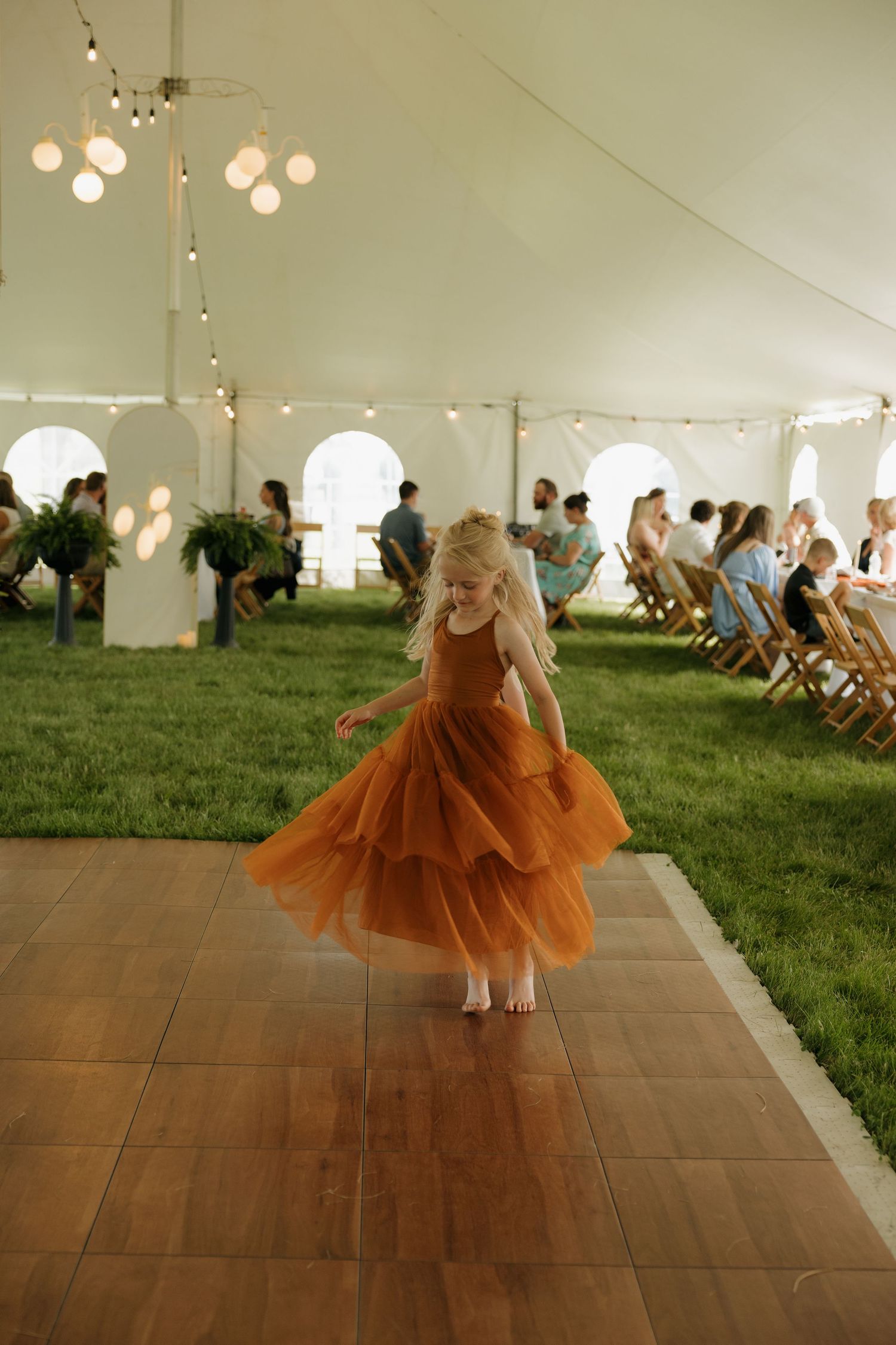 Young flower girl in orange tulle dress twirling barefoot on wooden dance floor under wedding tent.