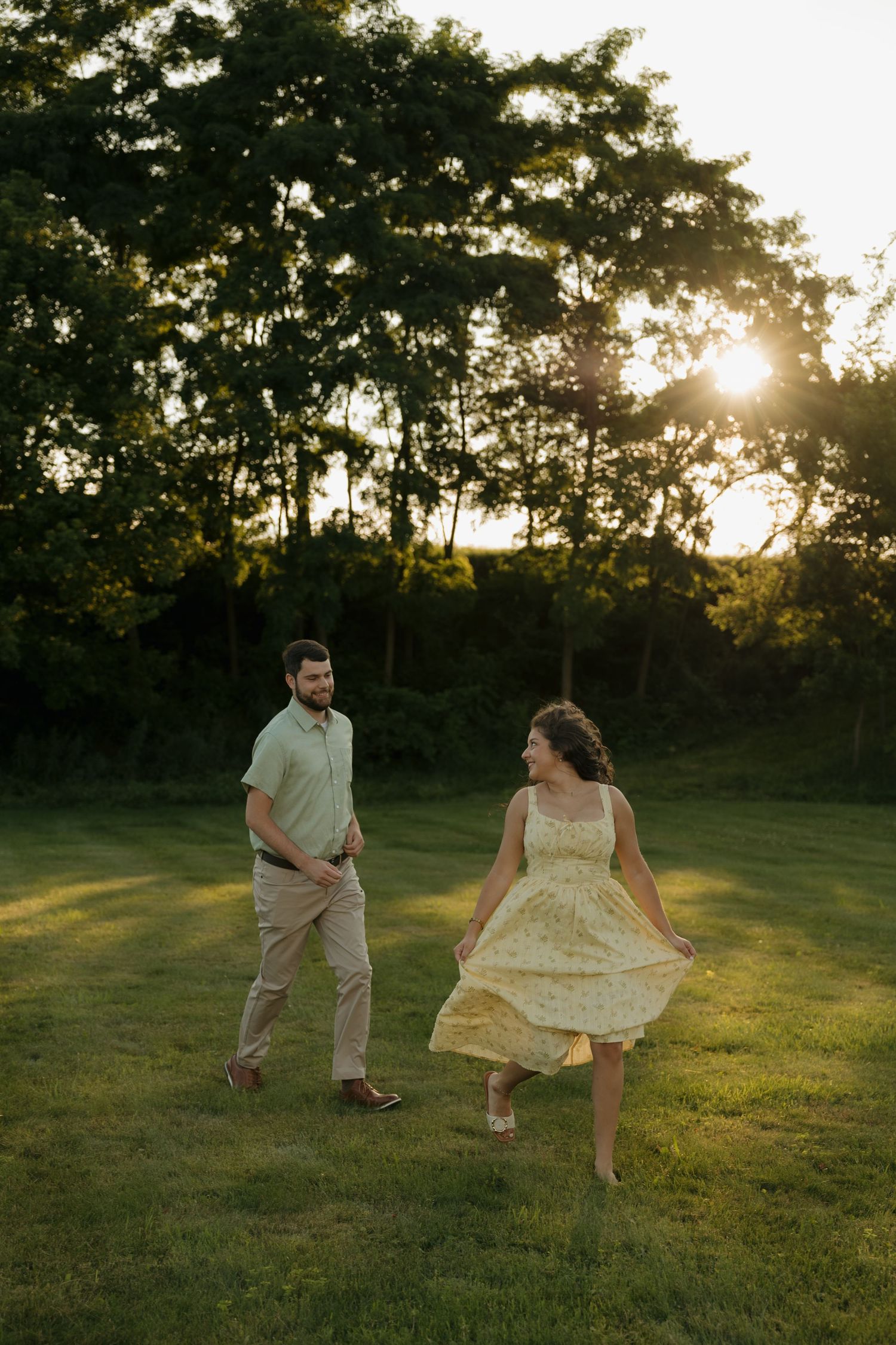 Couple dancing at golden hour sunset in lush green park, woman twirling yellow floral dress, man in sage green shirt smiling.