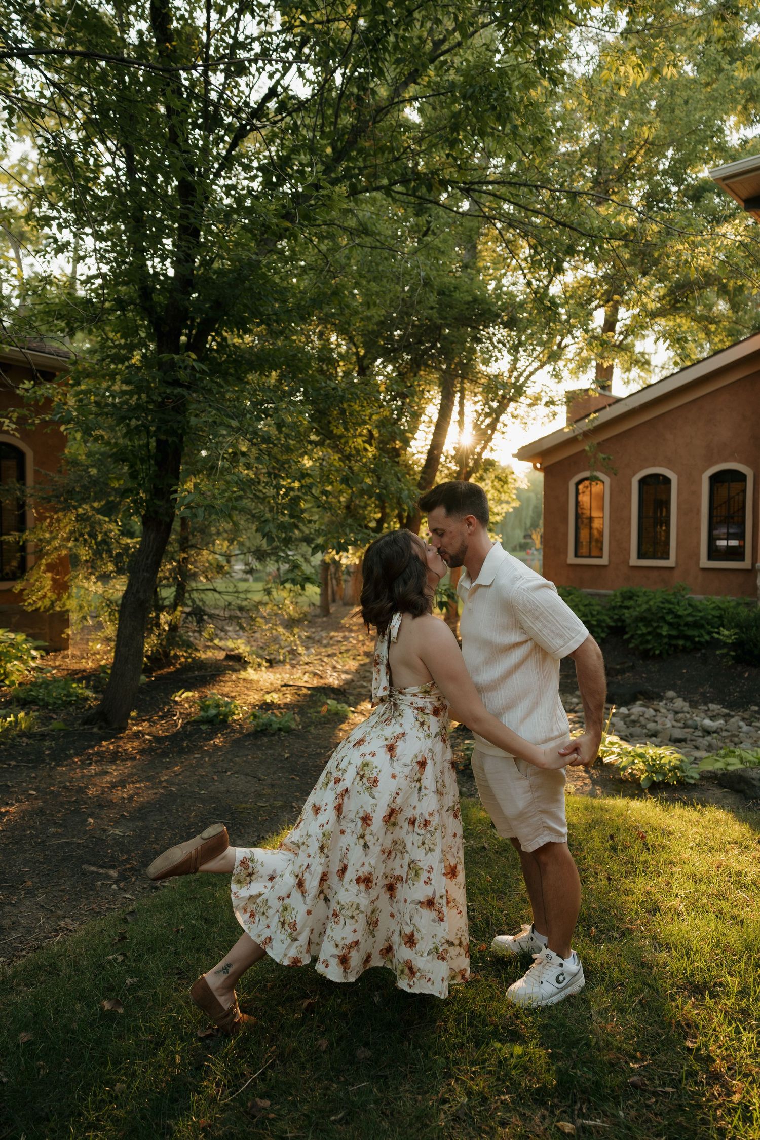 Couple sharing a romantic kiss while dancing in a sunlit garden near a brick building, woman wearing a floral dress.