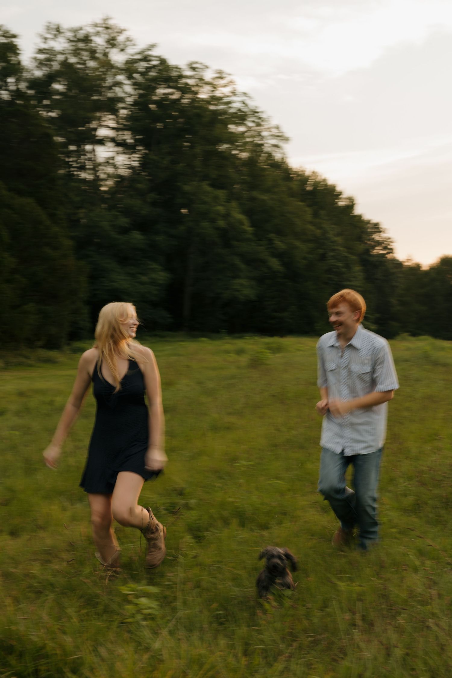A smiling couple runs through a lush green field with a small dog at dusk, trees in background.