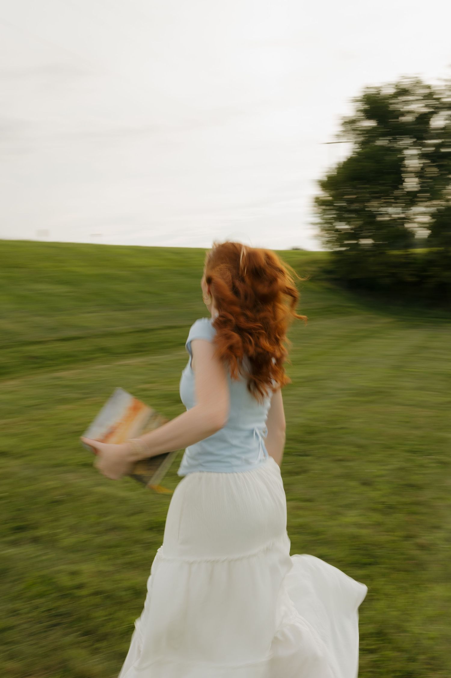 A redhead woman in white skirt and blue top runs through green field holding a book, motion blur effect.