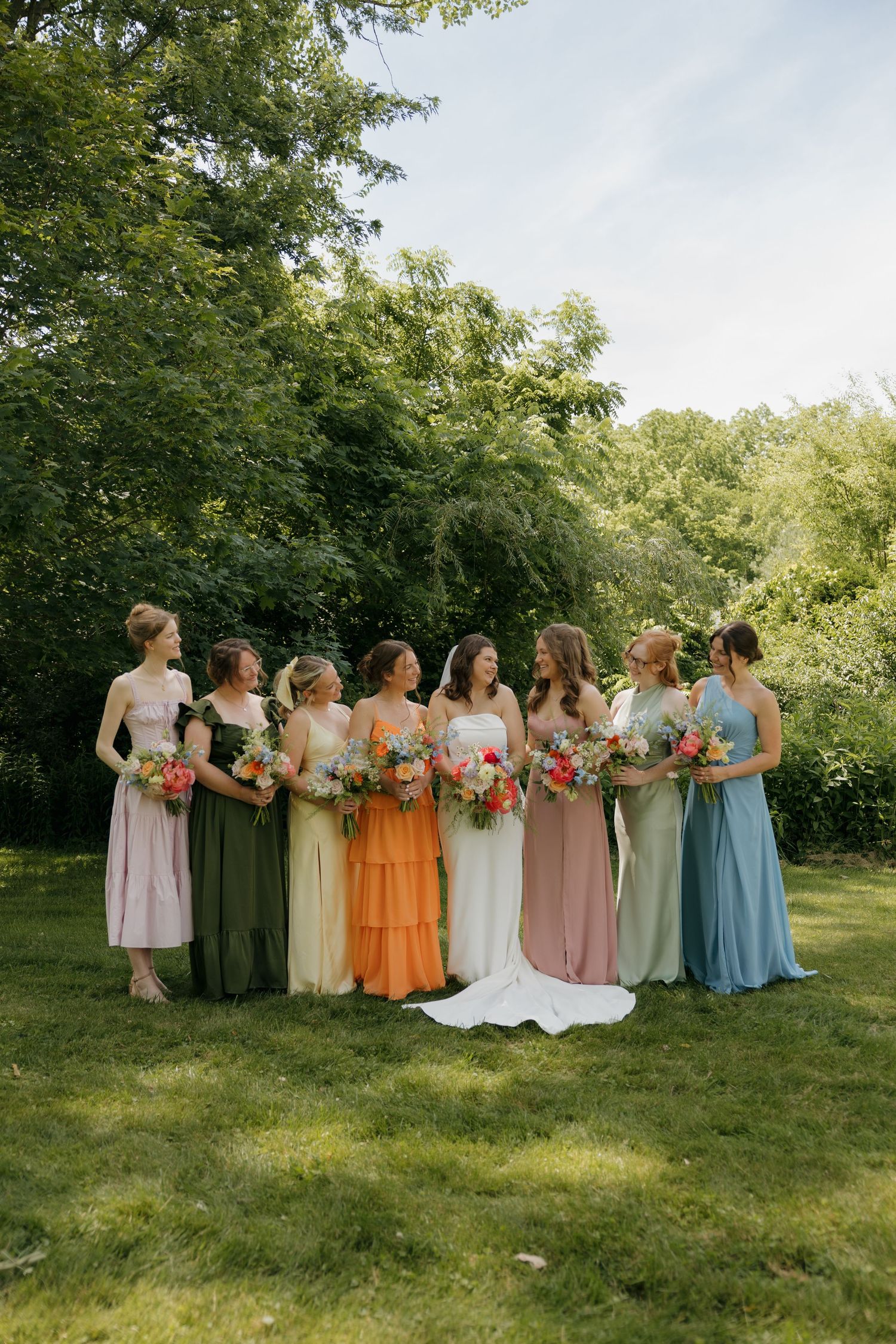 Bride and bridesmaids in mismatched colorful dresses holding floral bouquets standing outdoors on green grass.