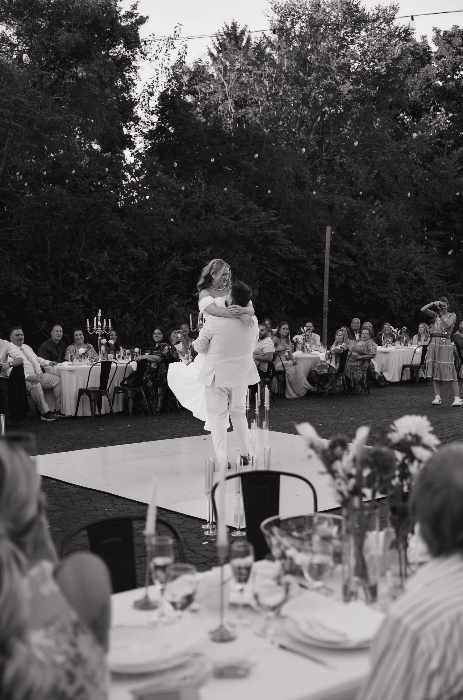 Black and white photo of groom lifting bride on outdoor dance floor surrounded by wedding guests at evening reception.