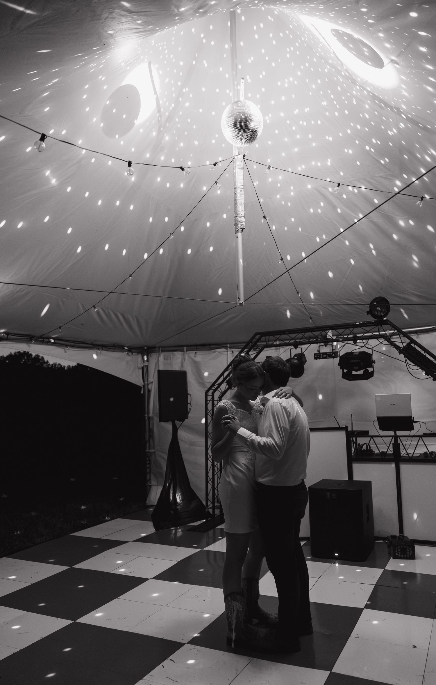Couple sharing first dance under disco ball in elegant tent venue with checkered dance floor, black and white photo.