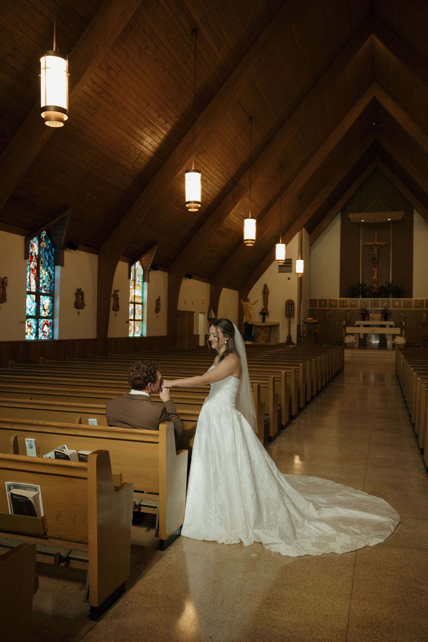 Bride in a white gown with a long train holds a seated groom's hand in the aisle of a church with wooden pews and stained glass.