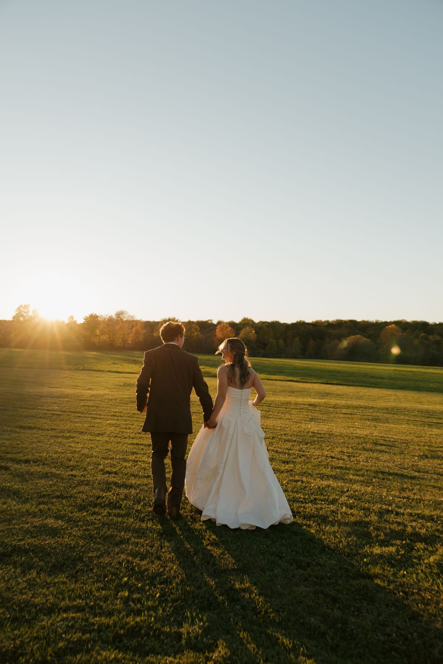 Bride in a white gown and groom in a dark suit hold hands walking across a grassy field toward a golden sunset.