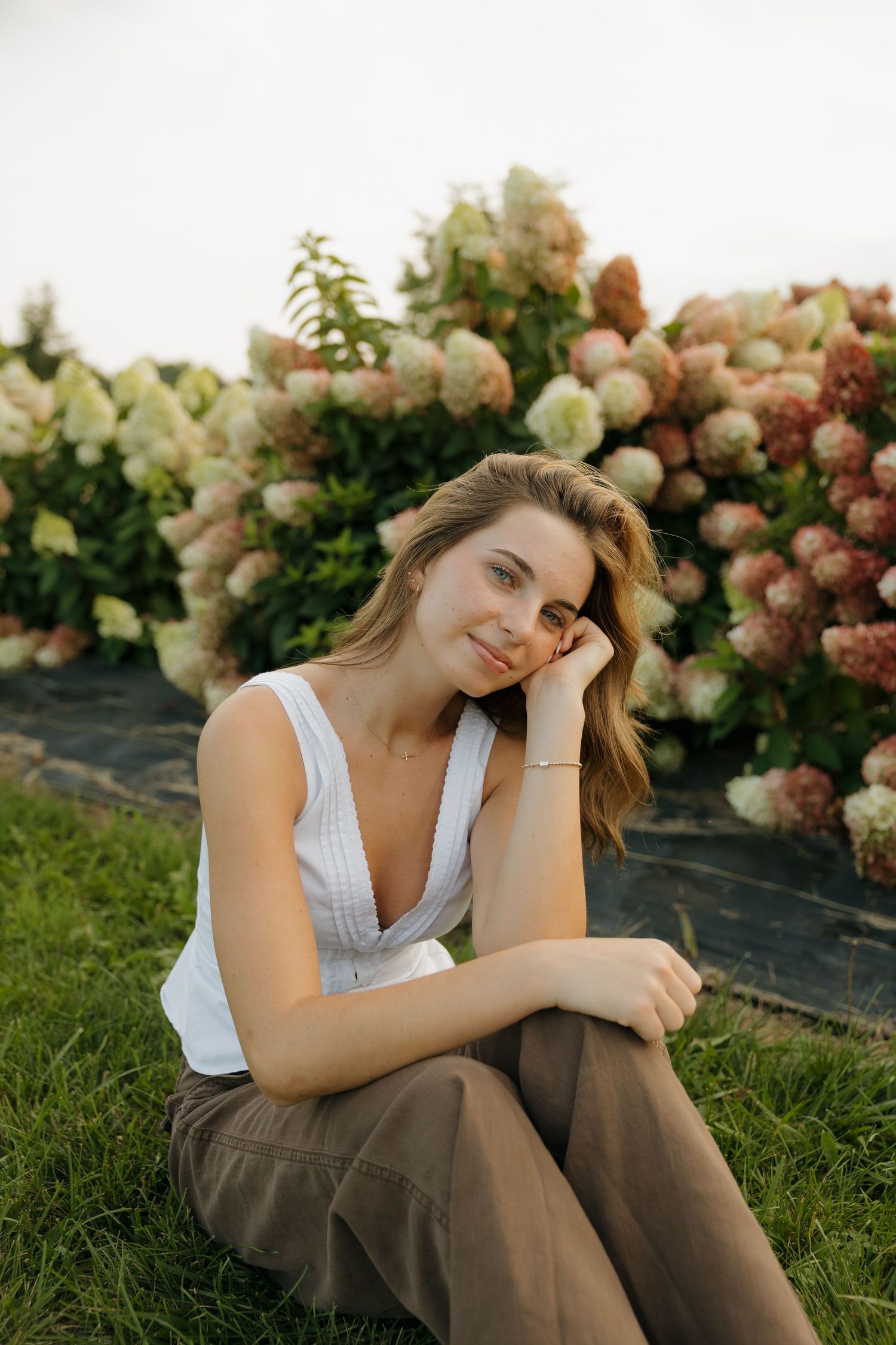 Young woman in white tank top and brown pants sitting on grass in front of blooming hydrangea bushes.