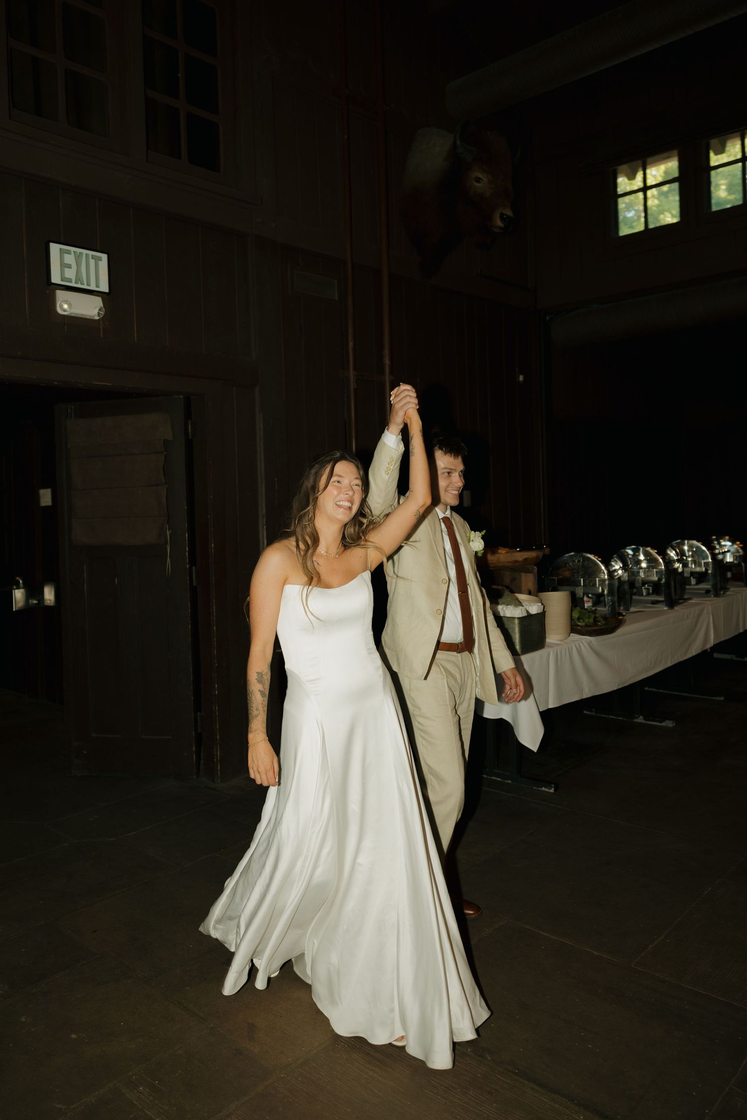 Bride in white satin gown and groom in tan suit share first dance, holding hands raised, in dimly lit venue.