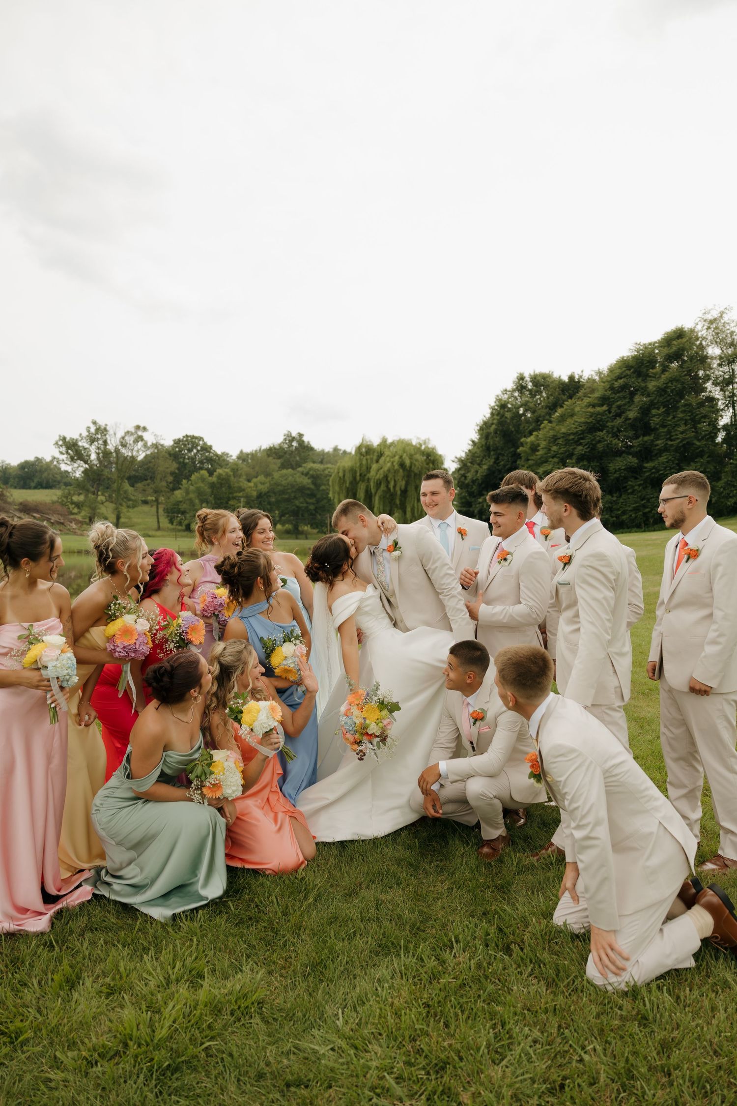 Wedding party poses on green lawn, colorful bridesmaid dresses contrast groomsmen in cream suits, bride and groom share a kiss.