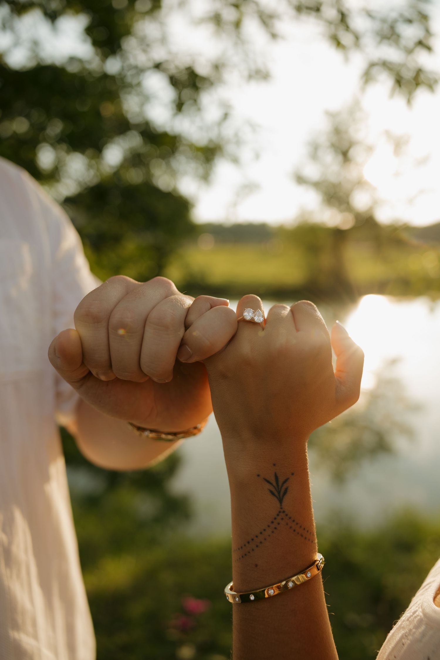 Two hands intertwined with an engagement ring and wrist tattoo, captured in golden sunset light near a serene lake.