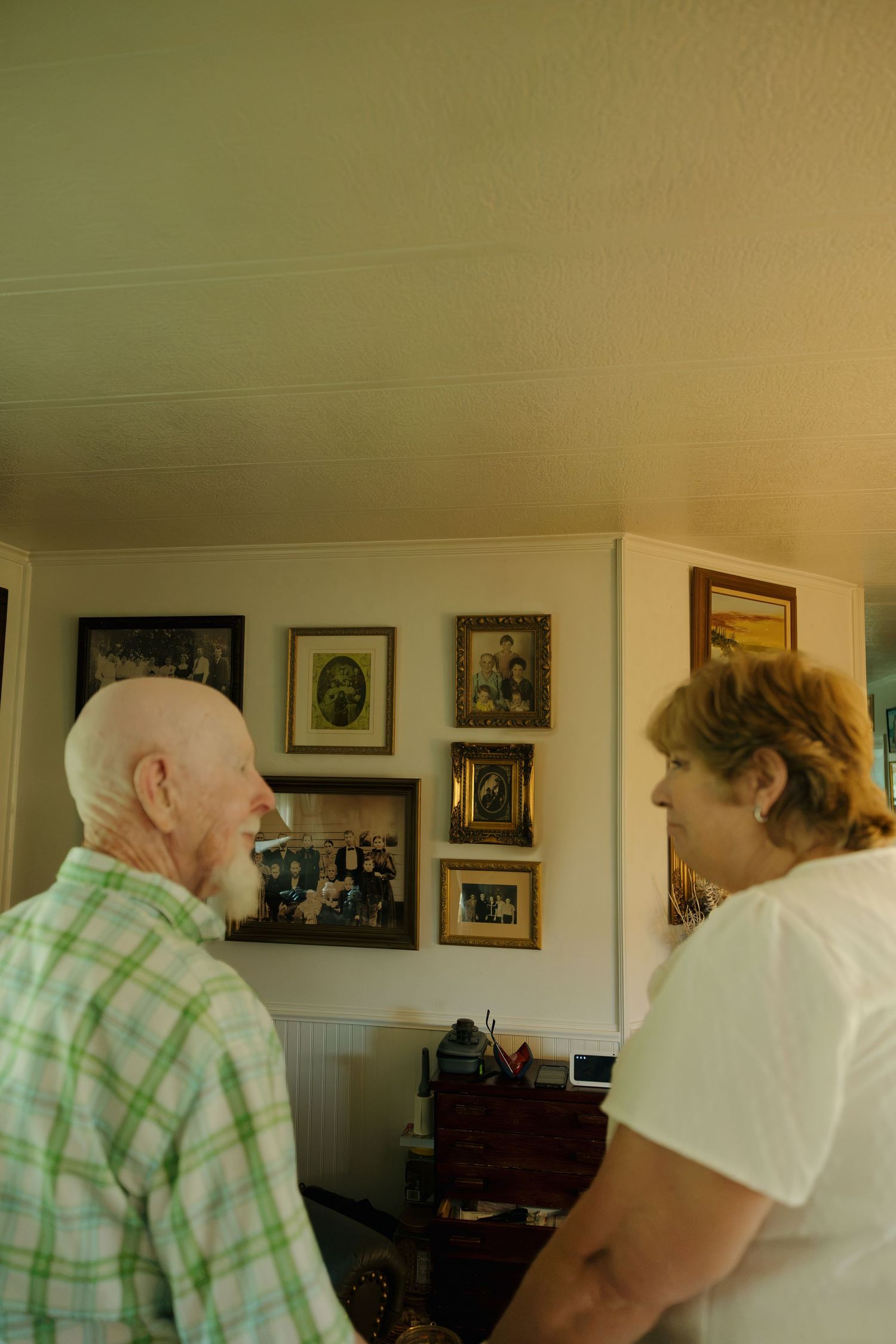 Elderly couple viewing framed family photos on a wall inside a cozy home.