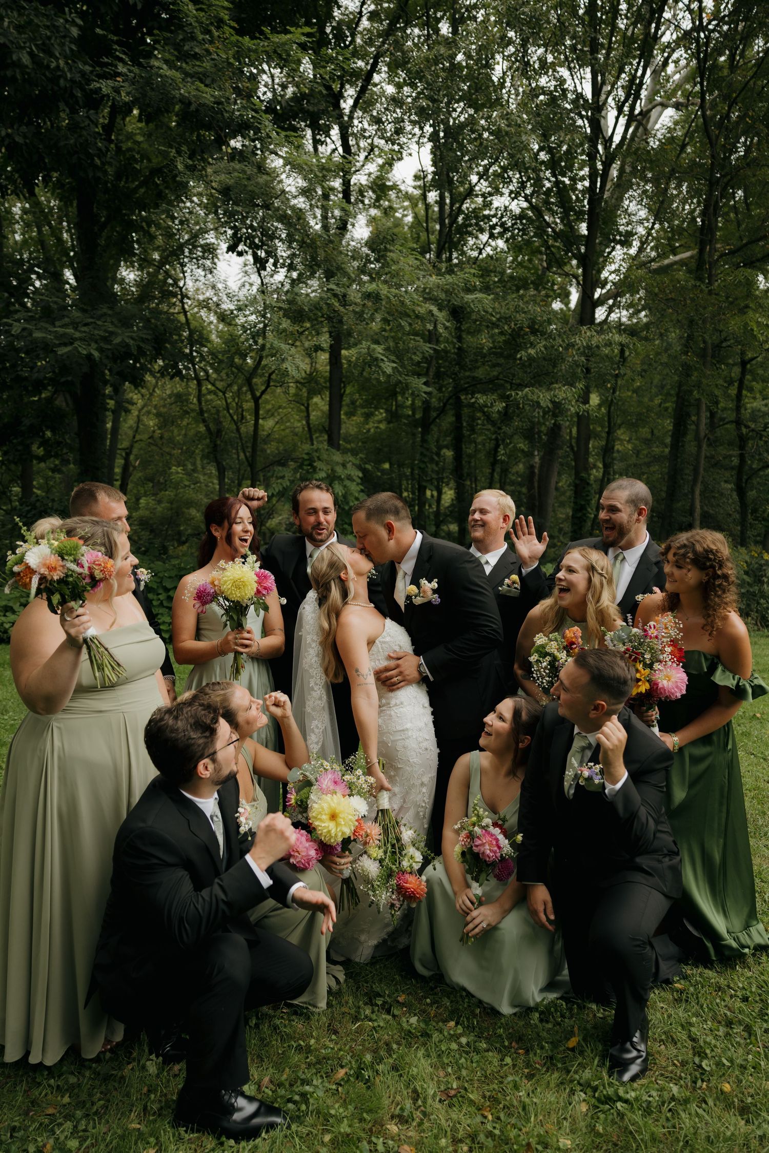 Wedding party in green dresses and black suits surrounds kissing bride and groom outdoors in lush forest setting.