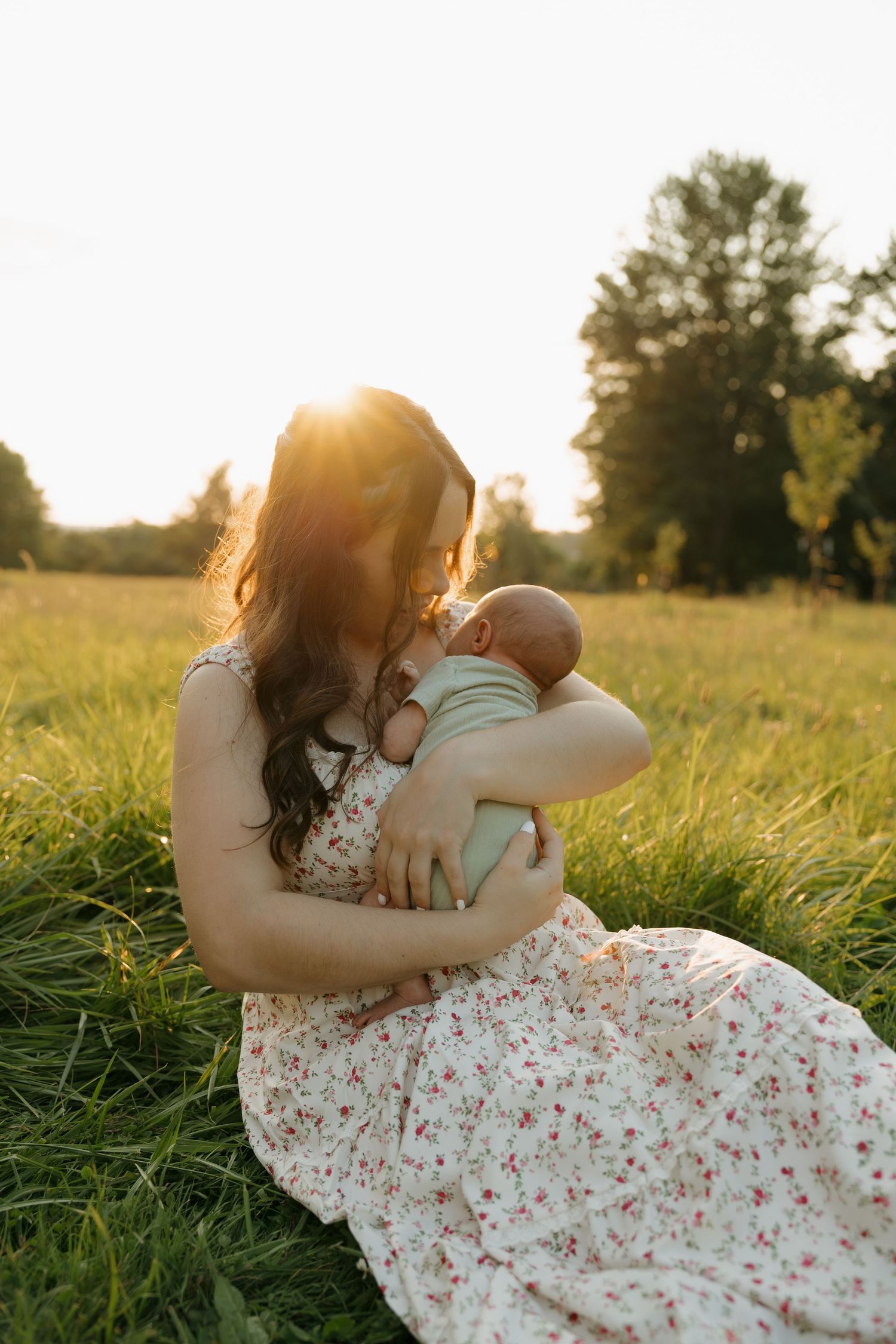 Mother in floral dress breastfeeding newborn baby while sitting in field at golden hour.