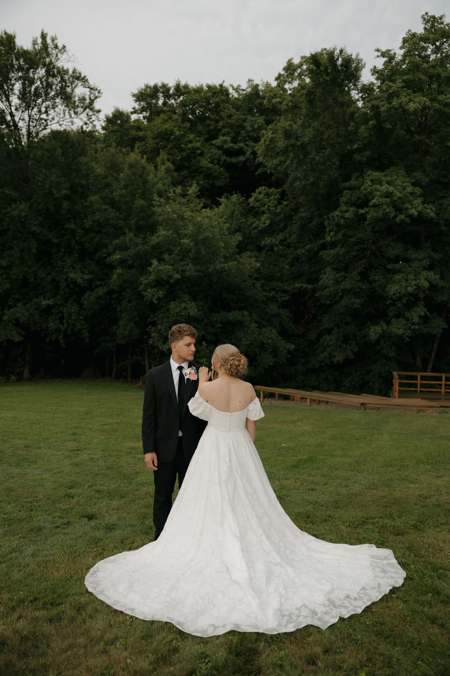 A bride in a white off-shoulder gown with a long train stands with a groom in a black tuxedo on a green lawn.