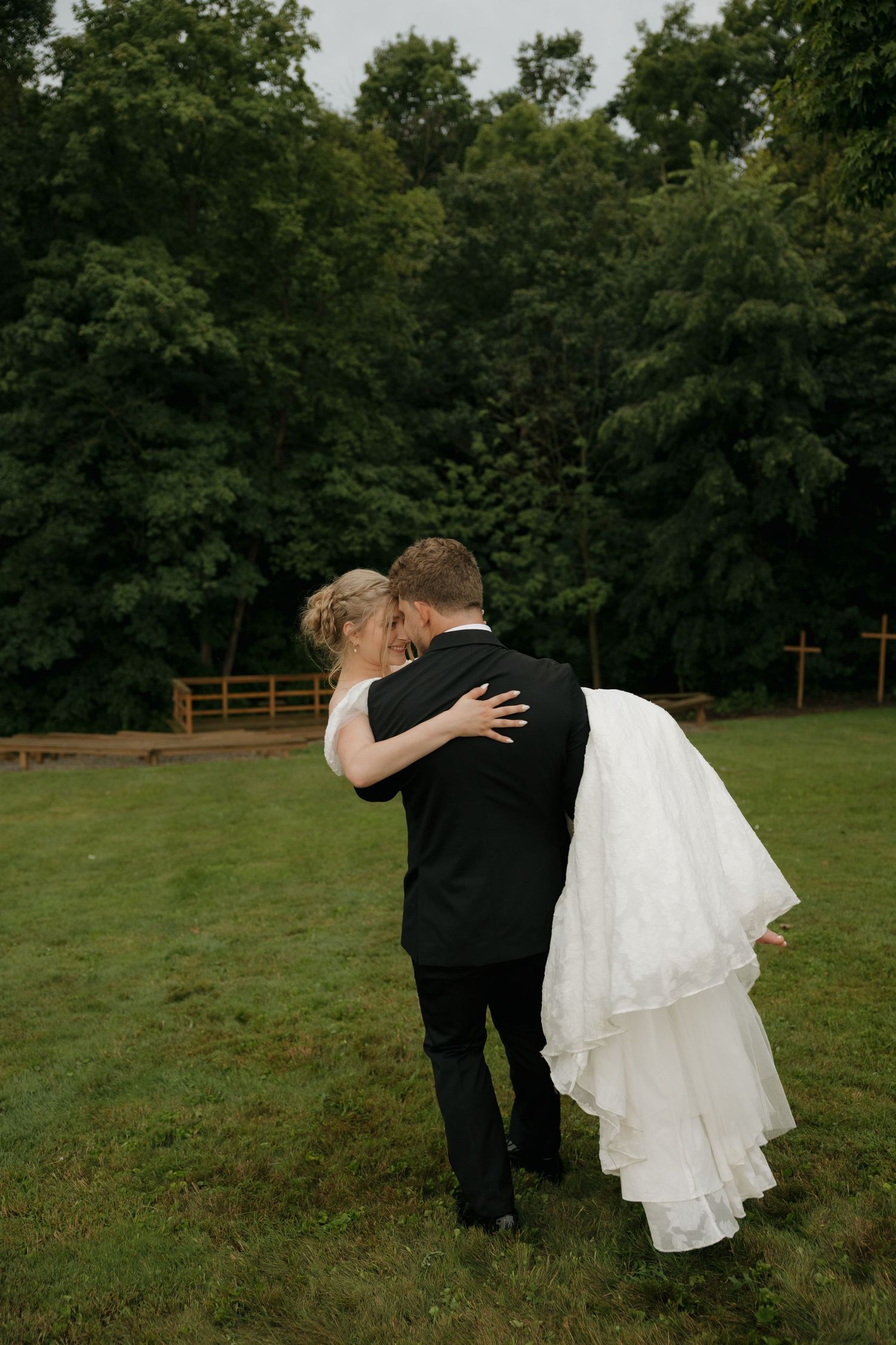 A bride in a white gown and groom in a black suit embrace on a green lawn with lush trees in the background.