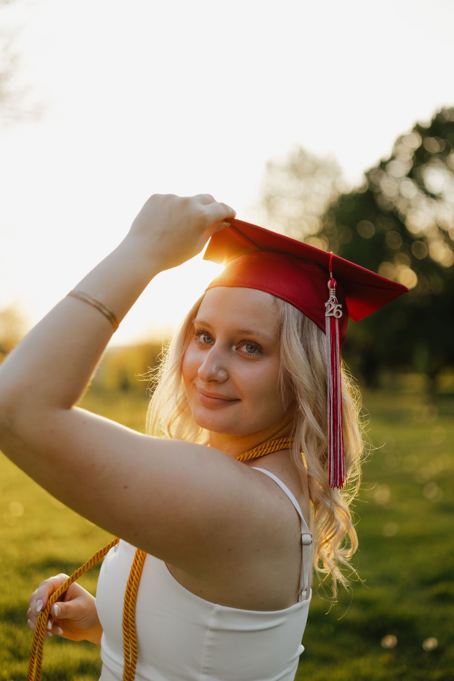 Blonde graduate holding red mortarboard cap with 25 tassel in golden sunset light outdoors.