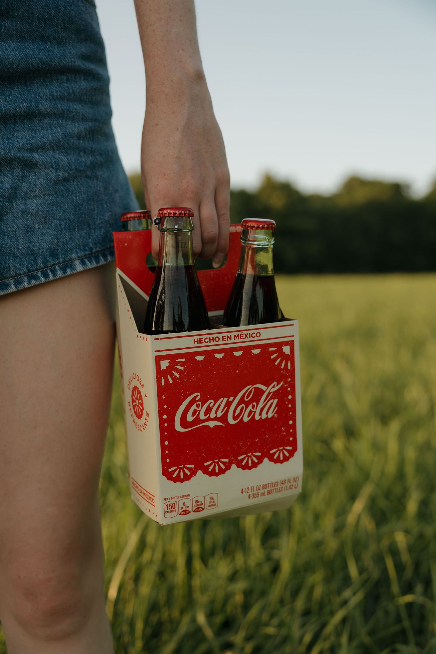 Person holding a 4-pack of Mexican Coca-Cola glass bottles in a cardboard carrier in a green field.