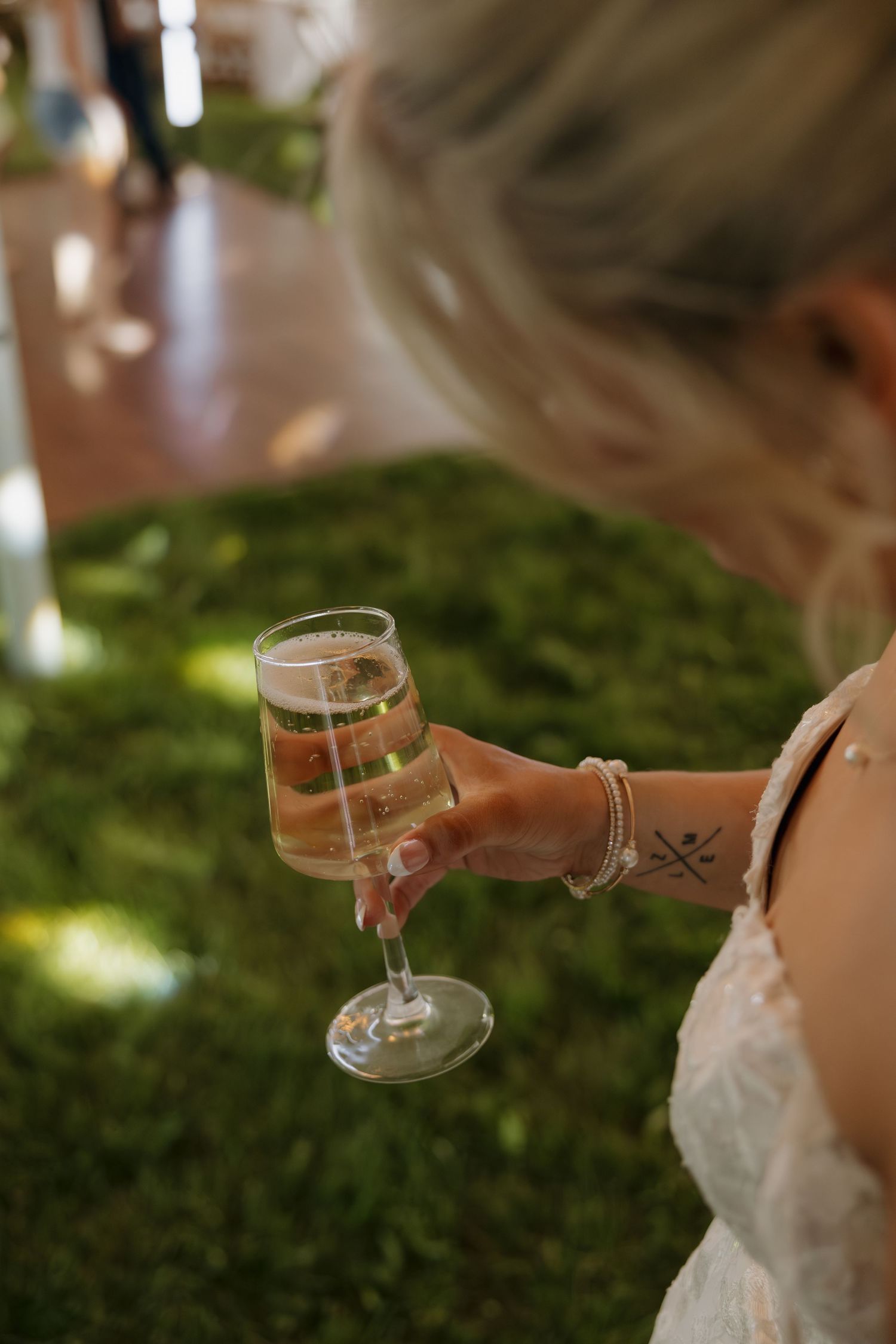 Blonde bride in lace dress holds champagne flute with tattoo and bracelets visible on wrist.