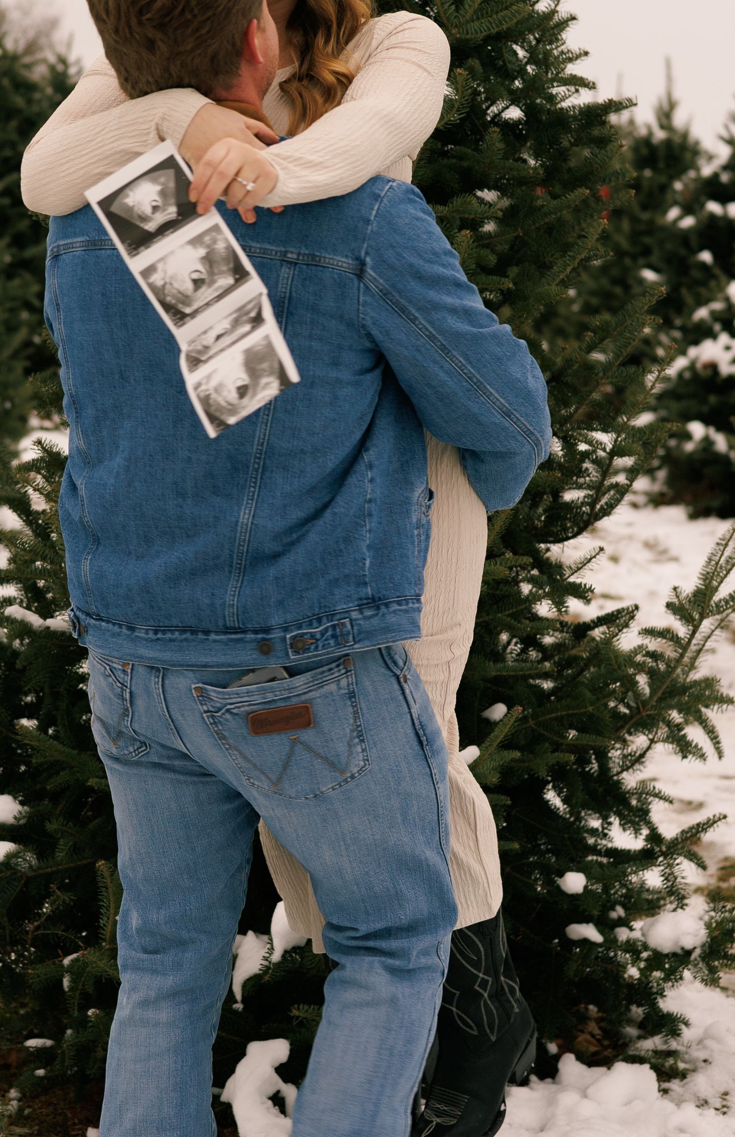 A couple hugs while holding a sonogram photo in front of snow-covered evergreen trees.