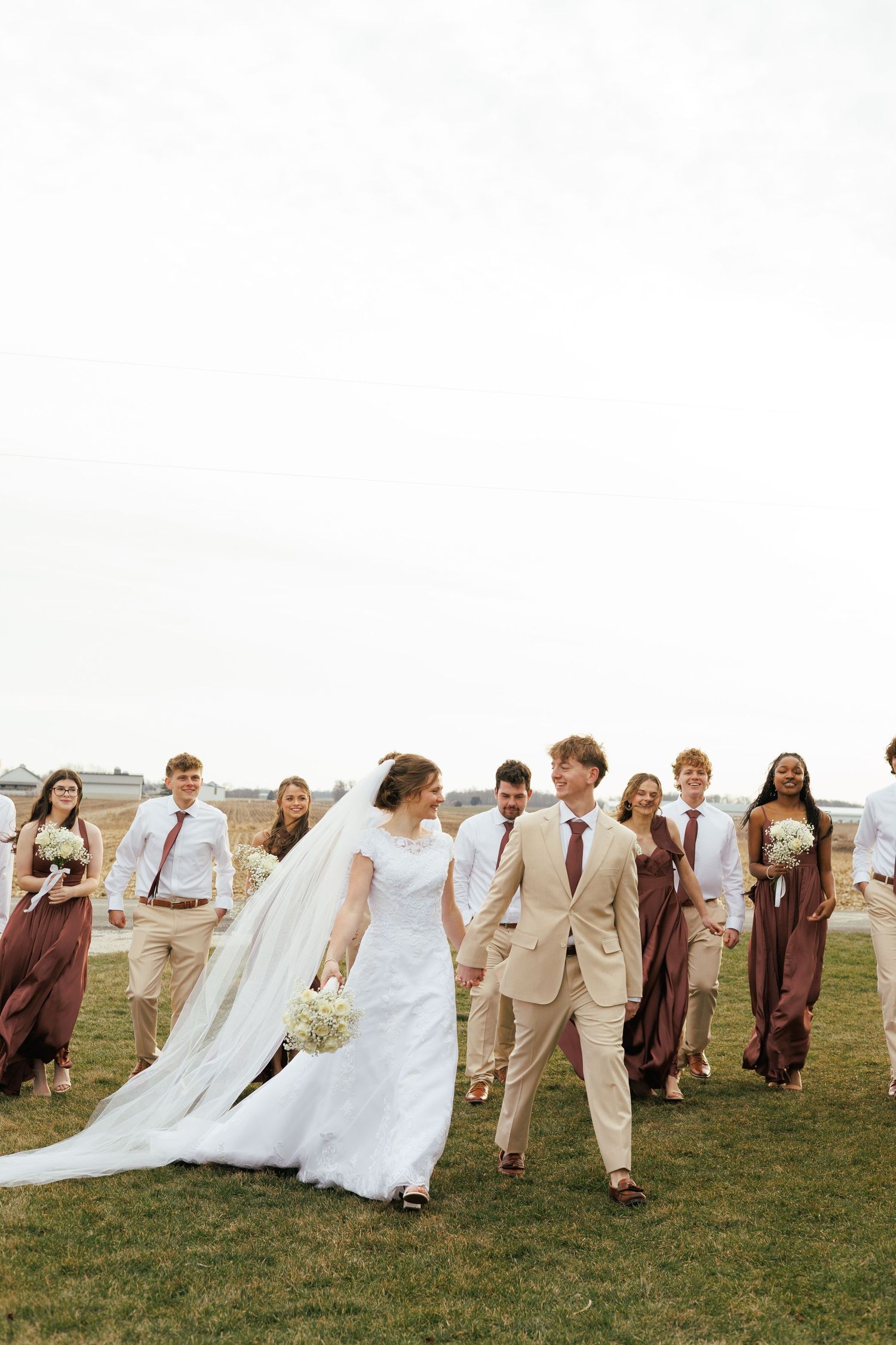 Wedding party walking and laughing together across a grassy field with flowing veils in the wind.