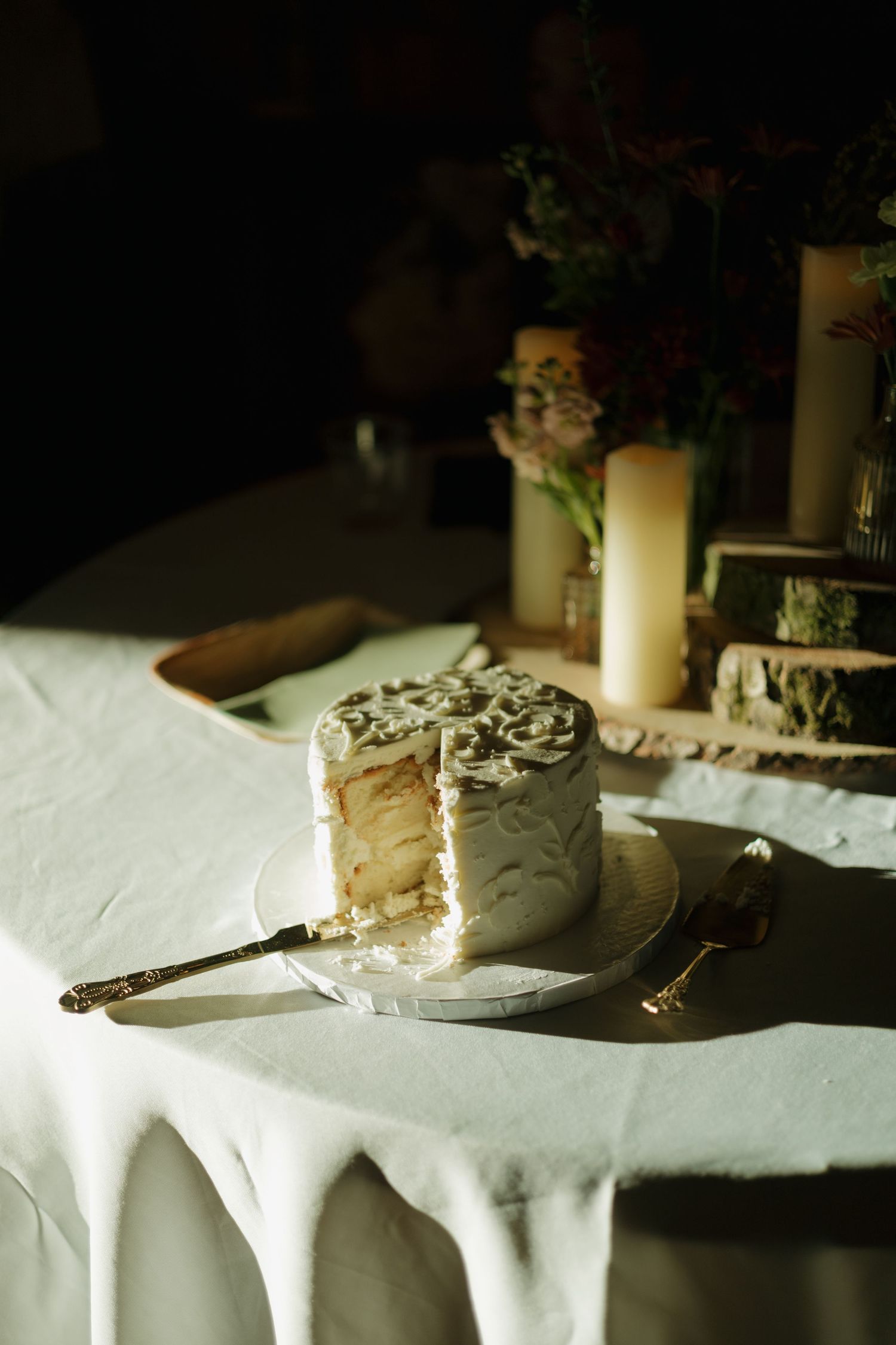 Partially sliced white frosted wedding cake with a gold-handled server on a draped table, beside pillar candles and rustic wood slices.