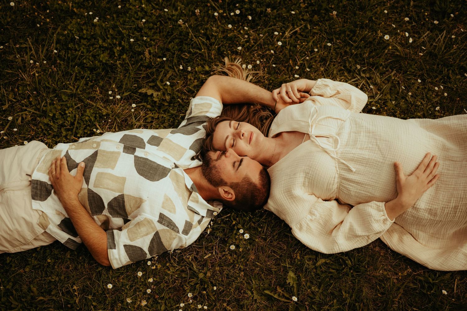 Photo romantique d'un couple de futurs parents allongés dans l'herbe, vêtus de blanc, lors d'une séance photo lifestyle en extérieur