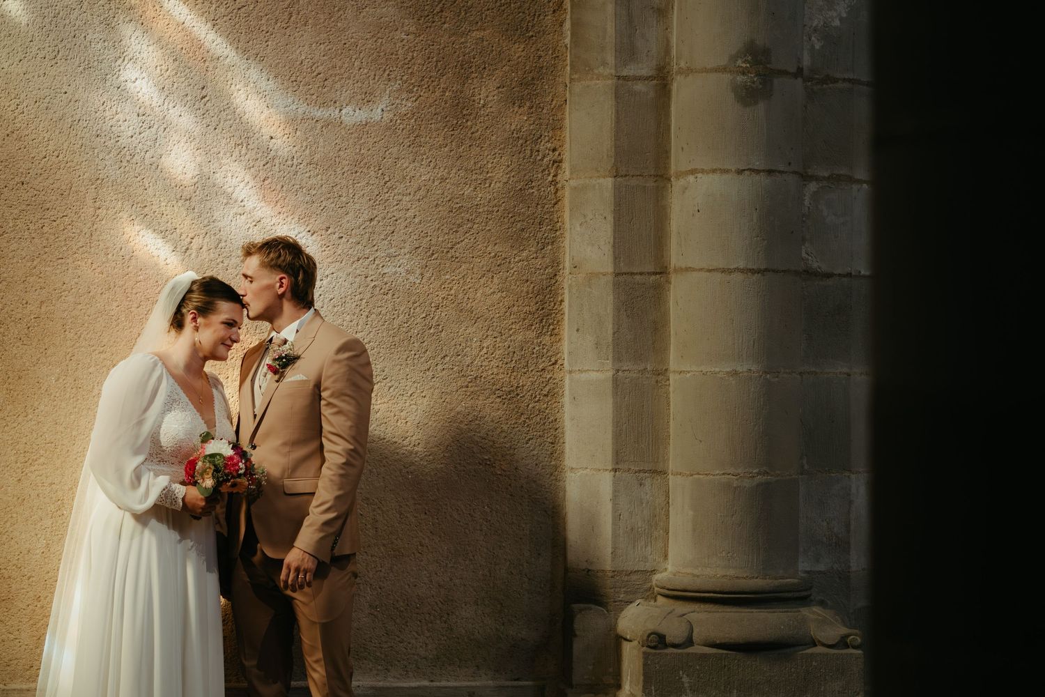 Mariée avec son voile, étant embrassée par son mari au costume marron, dans une église aux murs beiges, en Occitanie