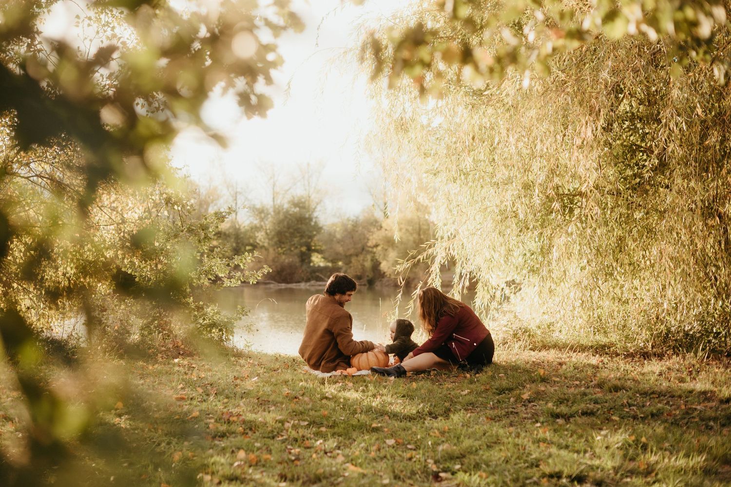Séance famille : des parents jouent avec leur enfant, assis dans l'herbe au coucher du soleil devant un lac à Bessières