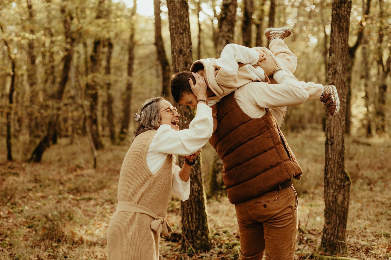 Séance famille : papa portant son fils et la maman riant, dans des tenues marrons et beige , en automne dans une forêt à Buzet sur Tarn