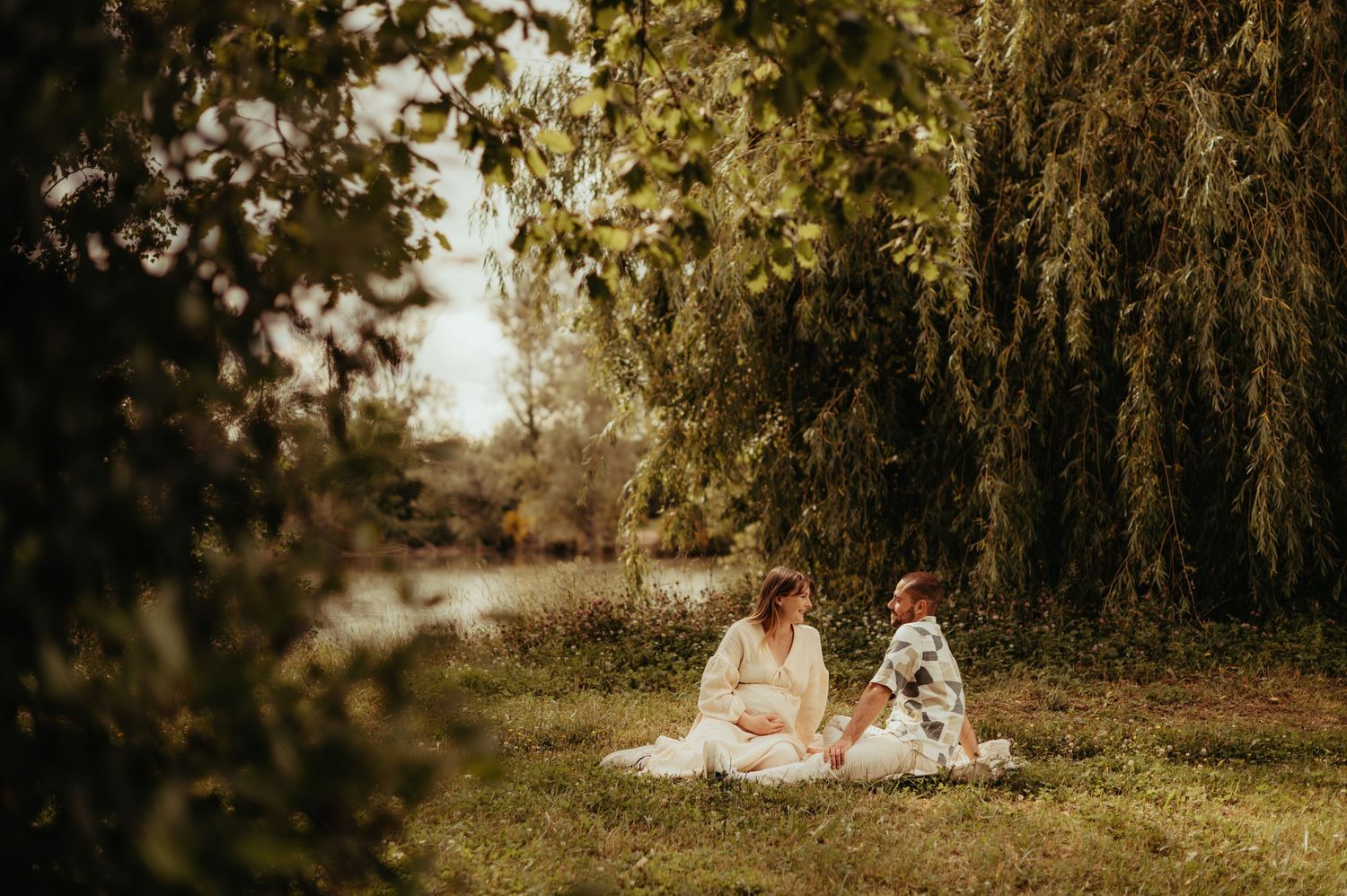 Un couple de futurs parents assis sur une couverture dans un champ doré au coucher du soleil, créant une scène automnale romantique.