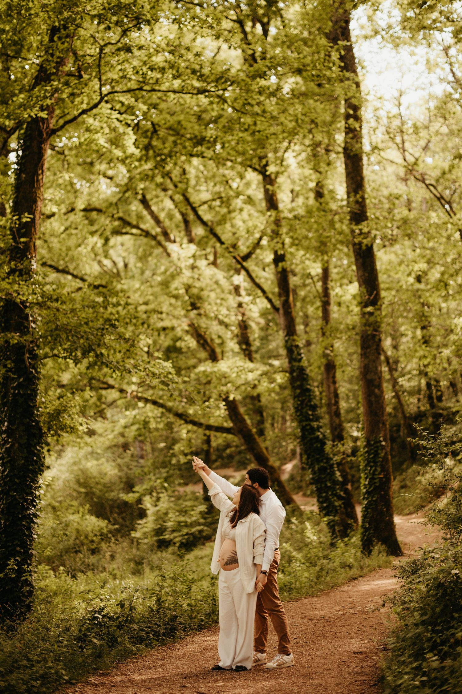 Séance maternité : future maman en tenue chemise ouverte beige, sur un sentier en forêt verdoyante, aux tons chauds et naturels dans le Tarn
