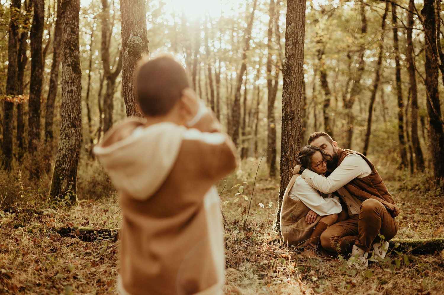 Séance famille, l'enfant regarde ses parents se câliner, en se bouchant les oreilles, dans une forêt automnale à Buzet sur Tarn