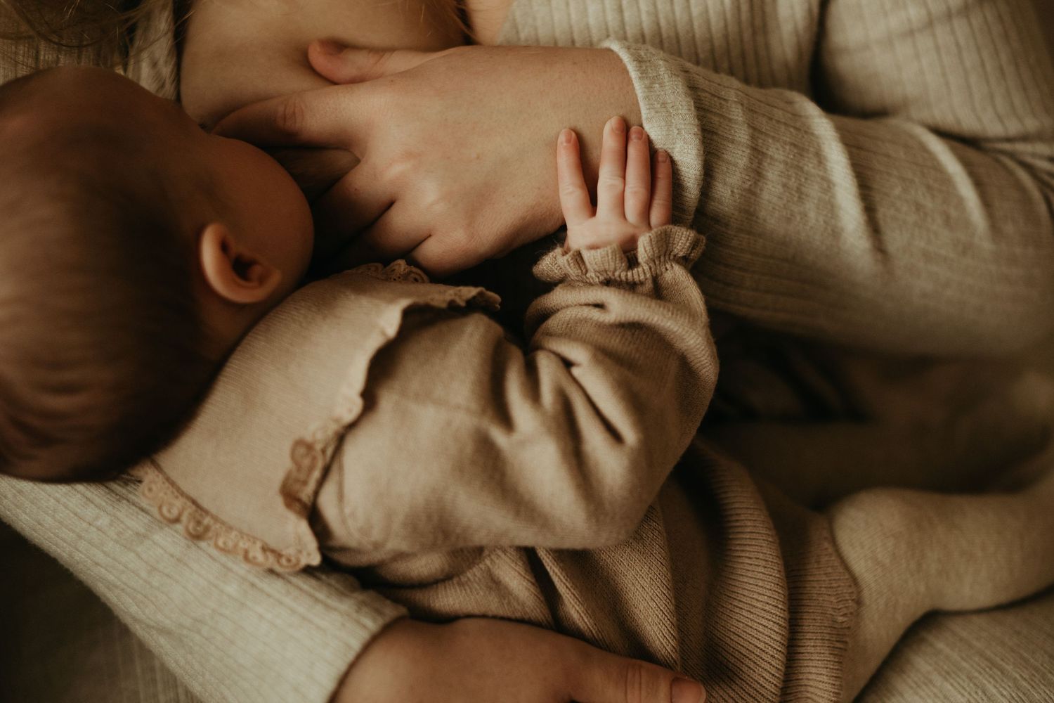 Séance nouveau-né : bébé tiens la main de sa maman pendant l'allaitement, dans des tenues beiges et tons chaud à Albi