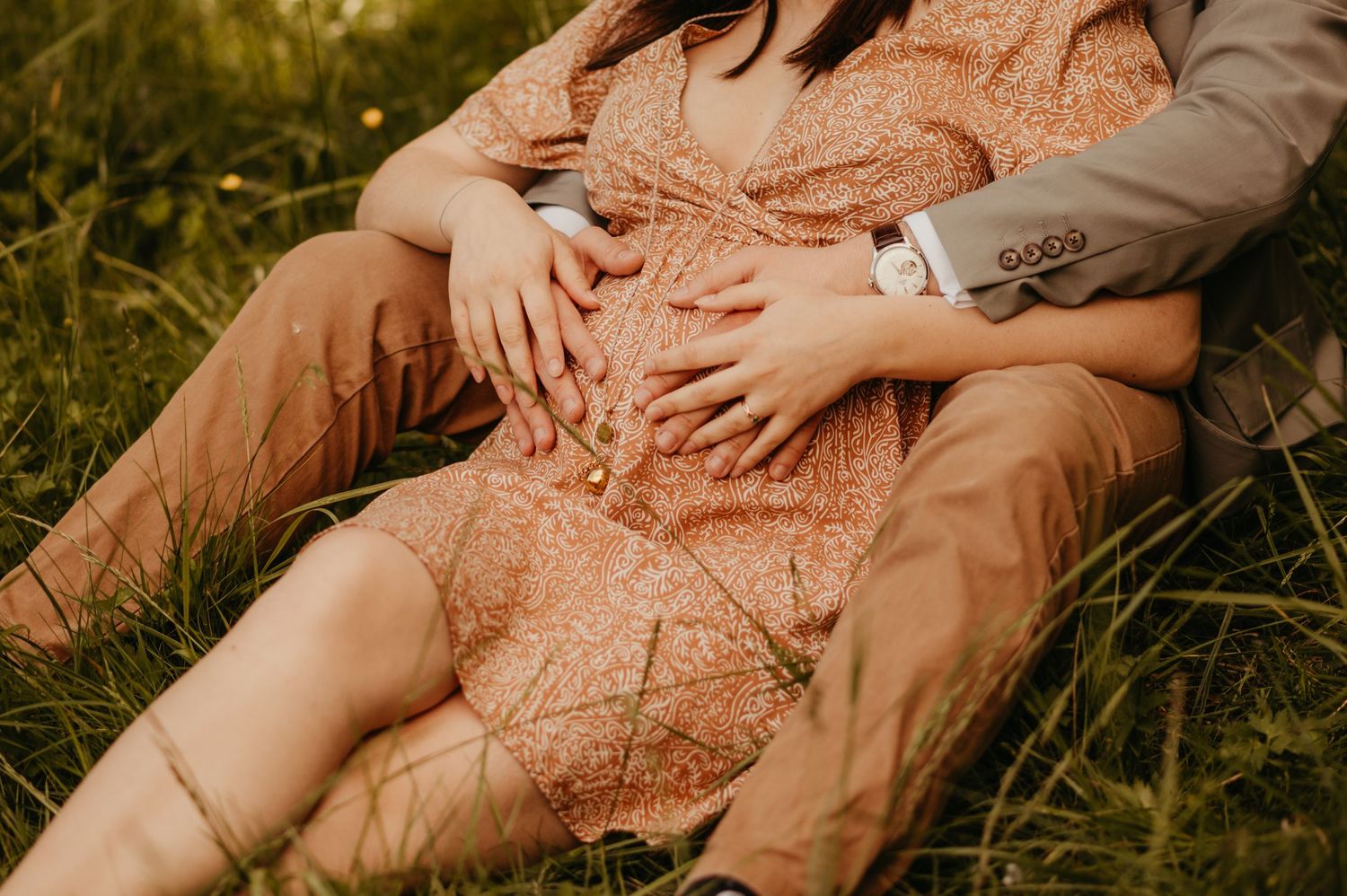 Une future maman vêtue d'une robe fluide couleur pêche est assise dans une prairie herbeuse à l'heure dorée, mains sur son ventre à Toulouse