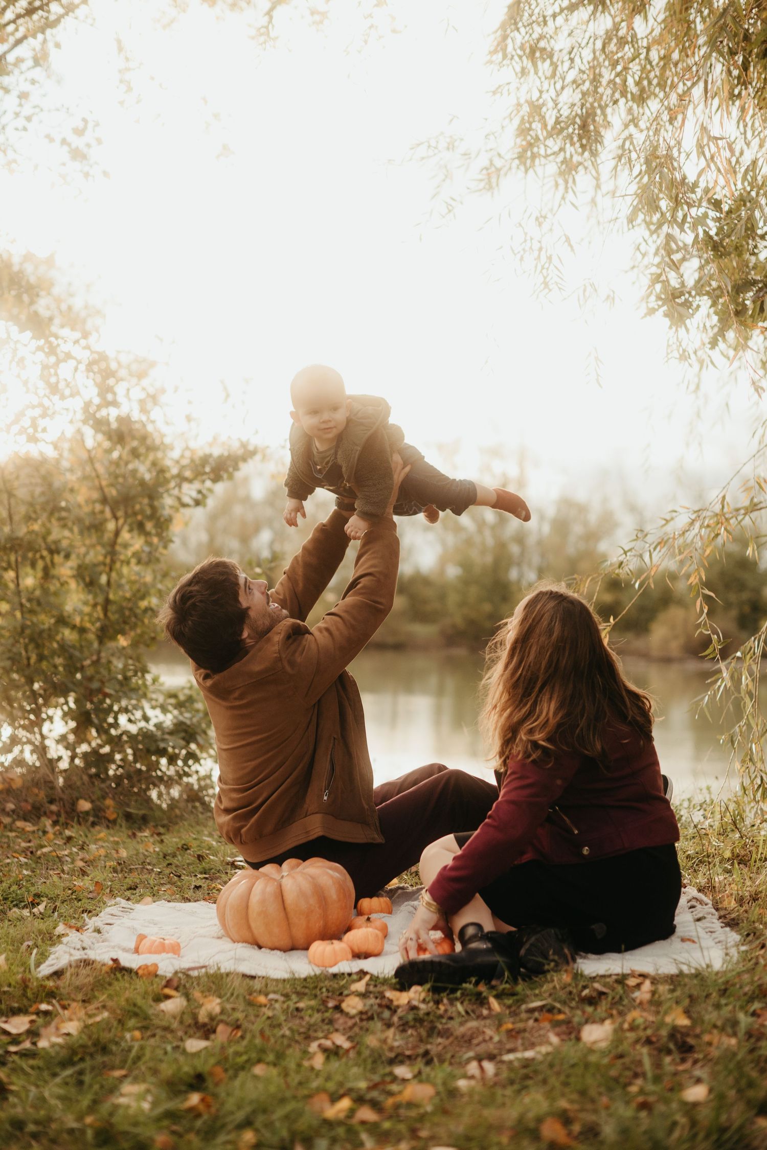 Papa qui tient son bébé, sous les yeux de maman en séance famille au bord d'un lac à Lisle sur Tarn
