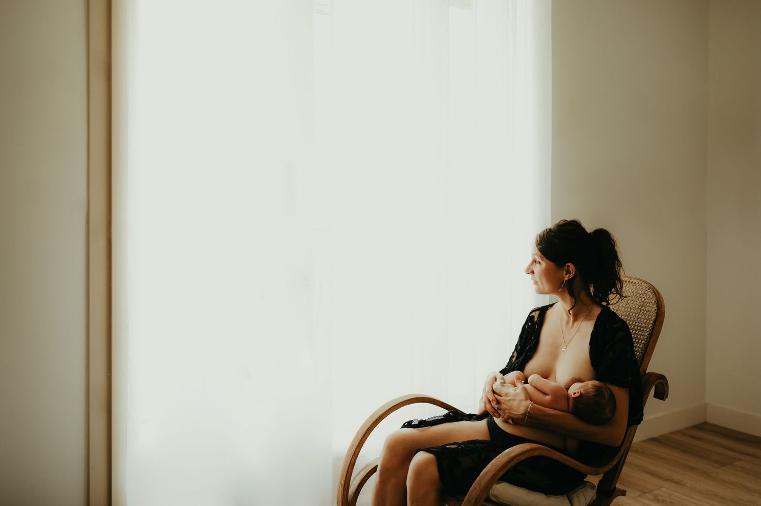 Bébé de 10 jours étant allaité par sa maman, qui regarde par la fenêtre, assise sur une chaise en rotin à Castres
