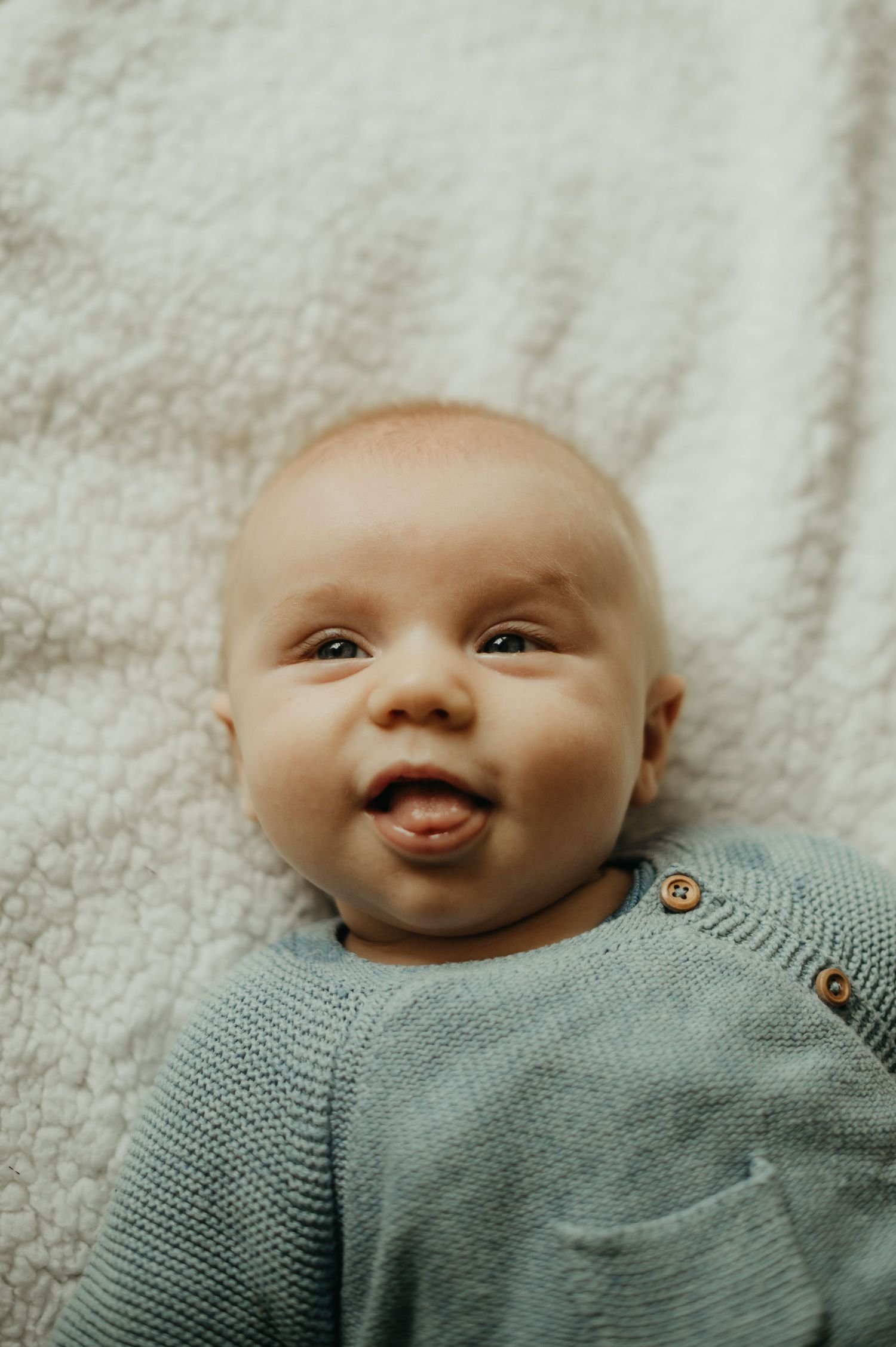 Un bébé souriant, vêtu d'un pull en maille, allongé sur une couverture couleur crème et affiche une expression heureuse en séance naissance